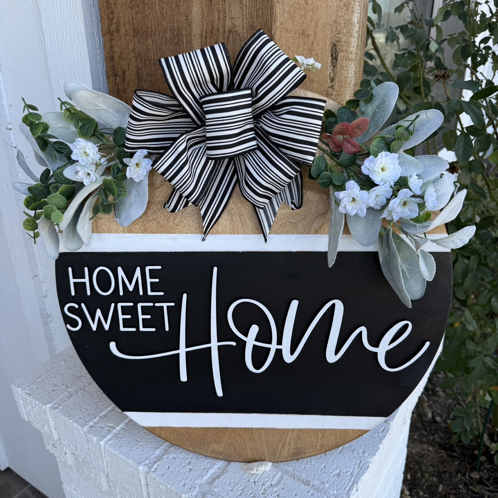 A decorative sign on a wood background reads Home Sweet Home in white script on a black semicircular board. It features a black and white striped bow at the top, with green foliage and white flowers on both sides, creating a welcoming and cozy aesthetic.