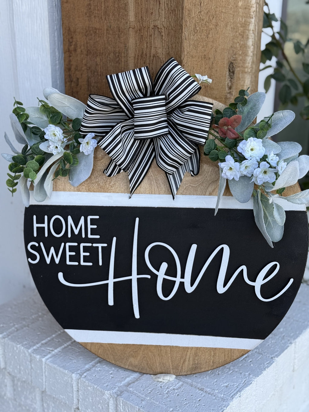A decorative sign reads Home Sweet Home in white script on a black background. It features a black and white striped bow at the top, flanked by green foliage and small white flowers, attached to a wooden base. The sign is propped against a wooden pillar with a white wall in the background.