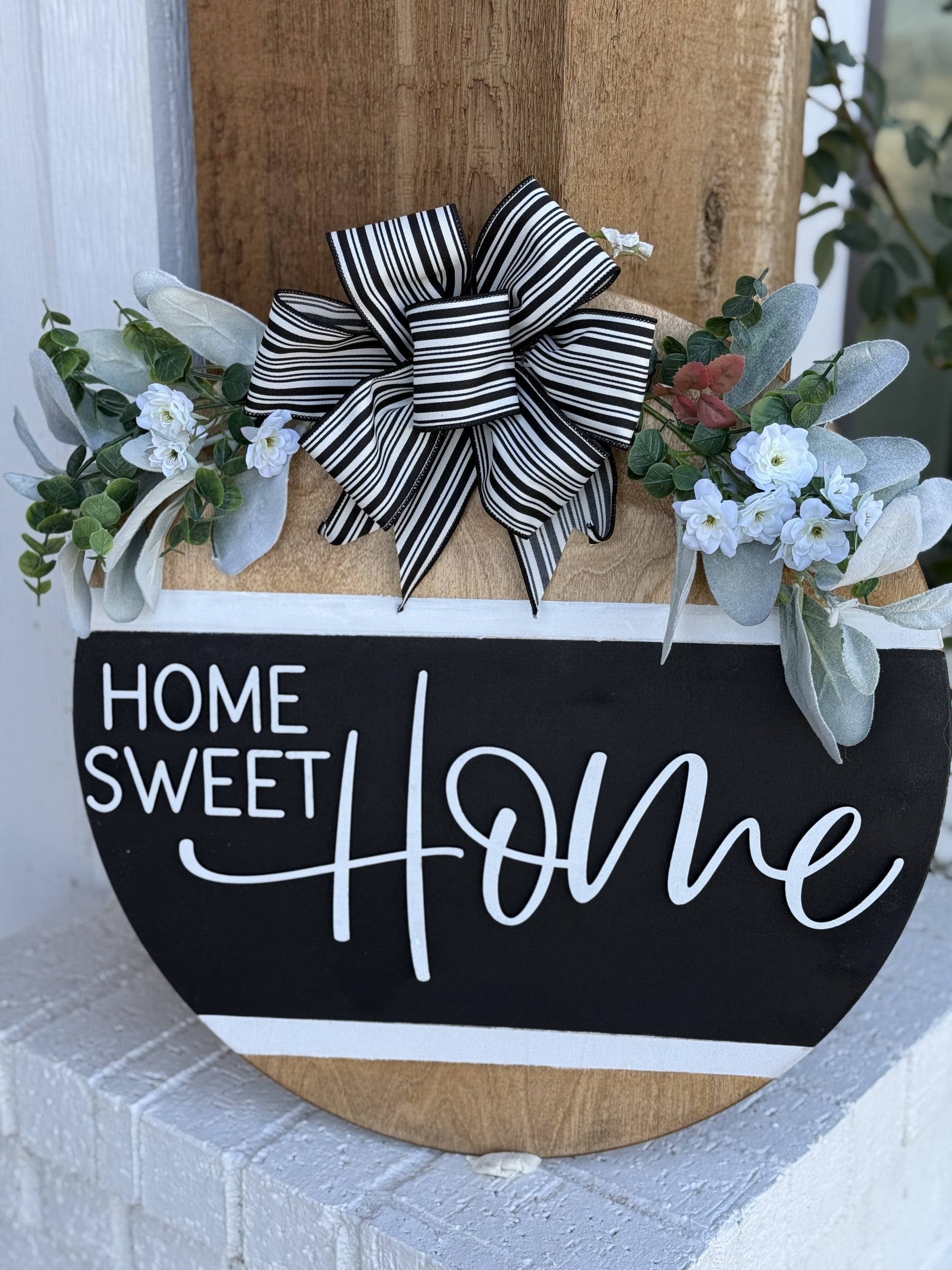 A decorative sign reads Home Sweet Home in white script on a black background. It features a black and white striped bow at the top, flanked by green foliage and small white flowers, attached to a wooden base. The sign is propped against a wooden pillar with a white wall in the background.