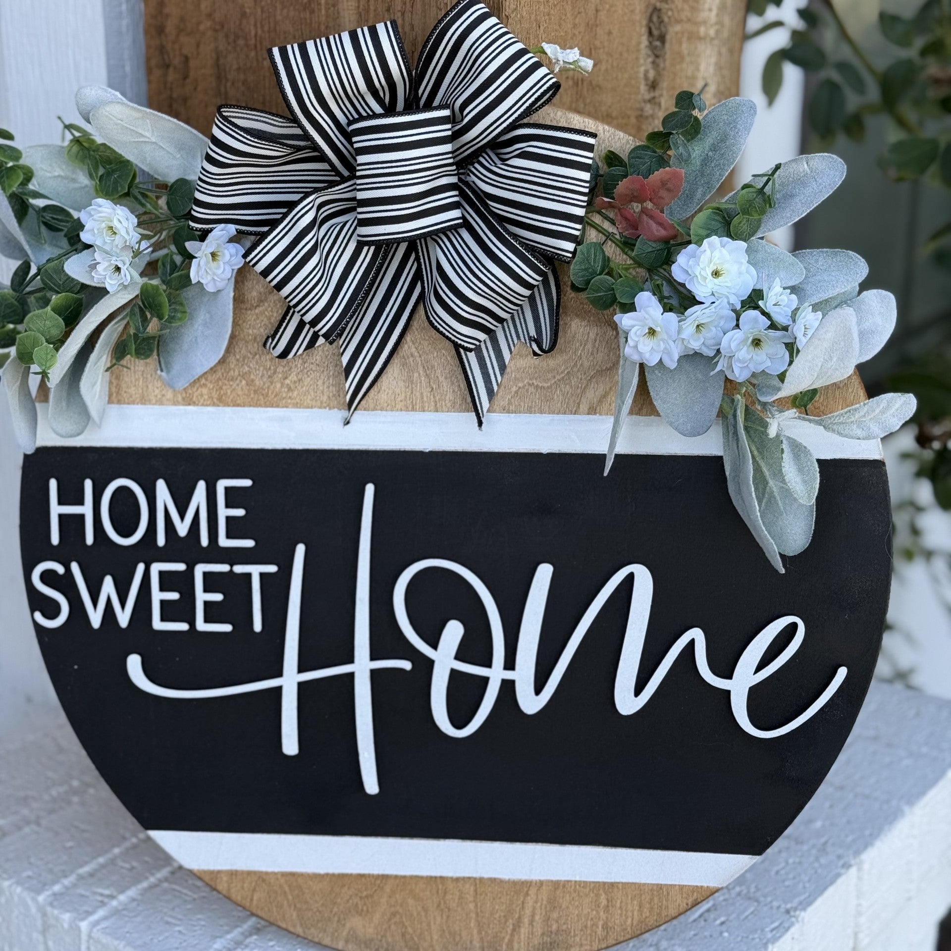 A wooden sign reads Home Sweet Home in white cursive on a black background. Its adorned with a large black and white striped bow at the top and flanked by white flowers and green leaves on both sides, creating a welcoming and cozy decoration.