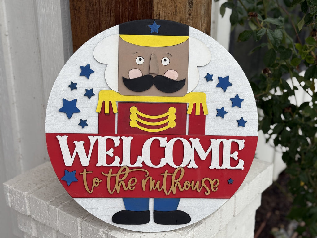 Round wooden sign with a Nutcracker soldier design. The soldier wears a yellow hat and black mustache. WELCOME to the nuthouse is written in bold white and gold letters. Blue stars surround the figure. The sign leans against a white brick surface and wooden background outdoors.