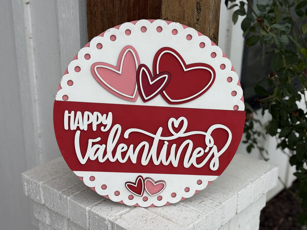 The Romantic Hearts Valentines Hanger features pink dots, Happy Valentines in white script on red, three hearts above (two pink, one red), and two smaller ones below. It hangs against a wooden post with green leaves, making it perfect for Valentines Day décor.