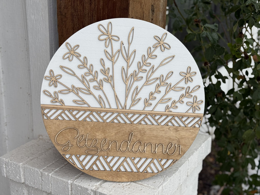 Round sign with floral design and the word Getendanner in the center. The top half features laser-cut flowers and leaves, while the bottom half has geometric patterns in natural wood tones. The sign rests on a white brick surface near greenery.