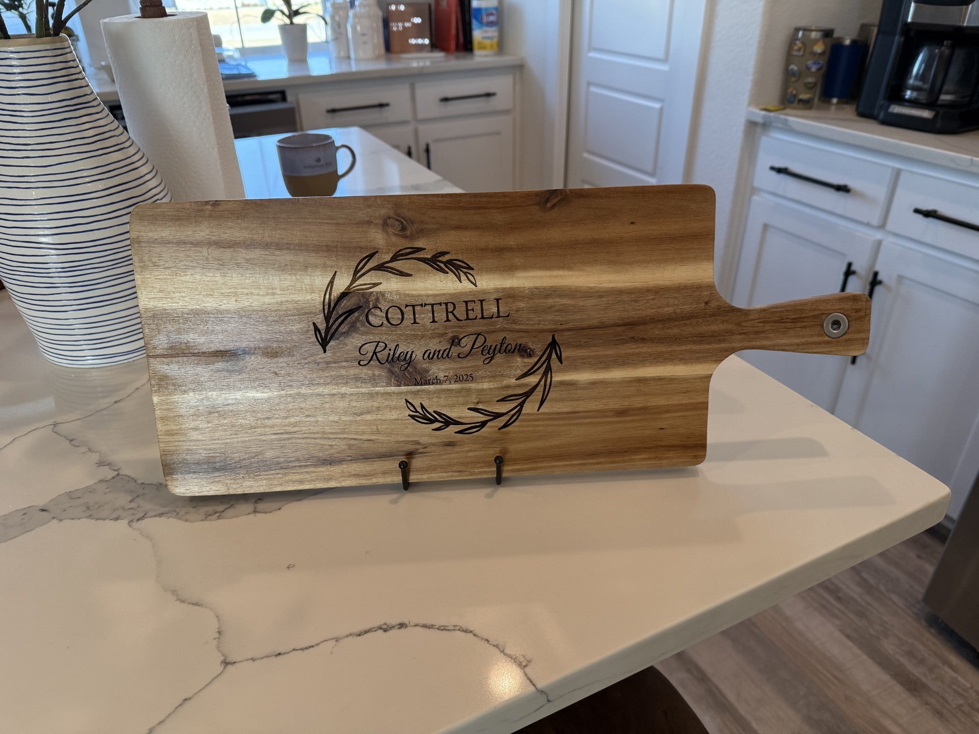 A personalized wooden cutting board, titled Personalized family information cutting board, stands on a kitchen counter with an engraved design featuring the name Cottrell and Riley and Peyton inside a decorative laurel wreath. The marble surface accents cabinets and appliances in the background.