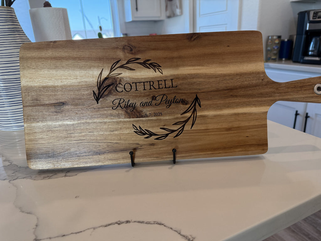 A personalized wooden cutting board, featuring an engraved wreath and Cottrell, Riley and Peyton, Est. March 8, 2023, with a hanging handle is displayed upright on a metal stand against the white marble kitchen counter. Product: Personalized family information cutting board.