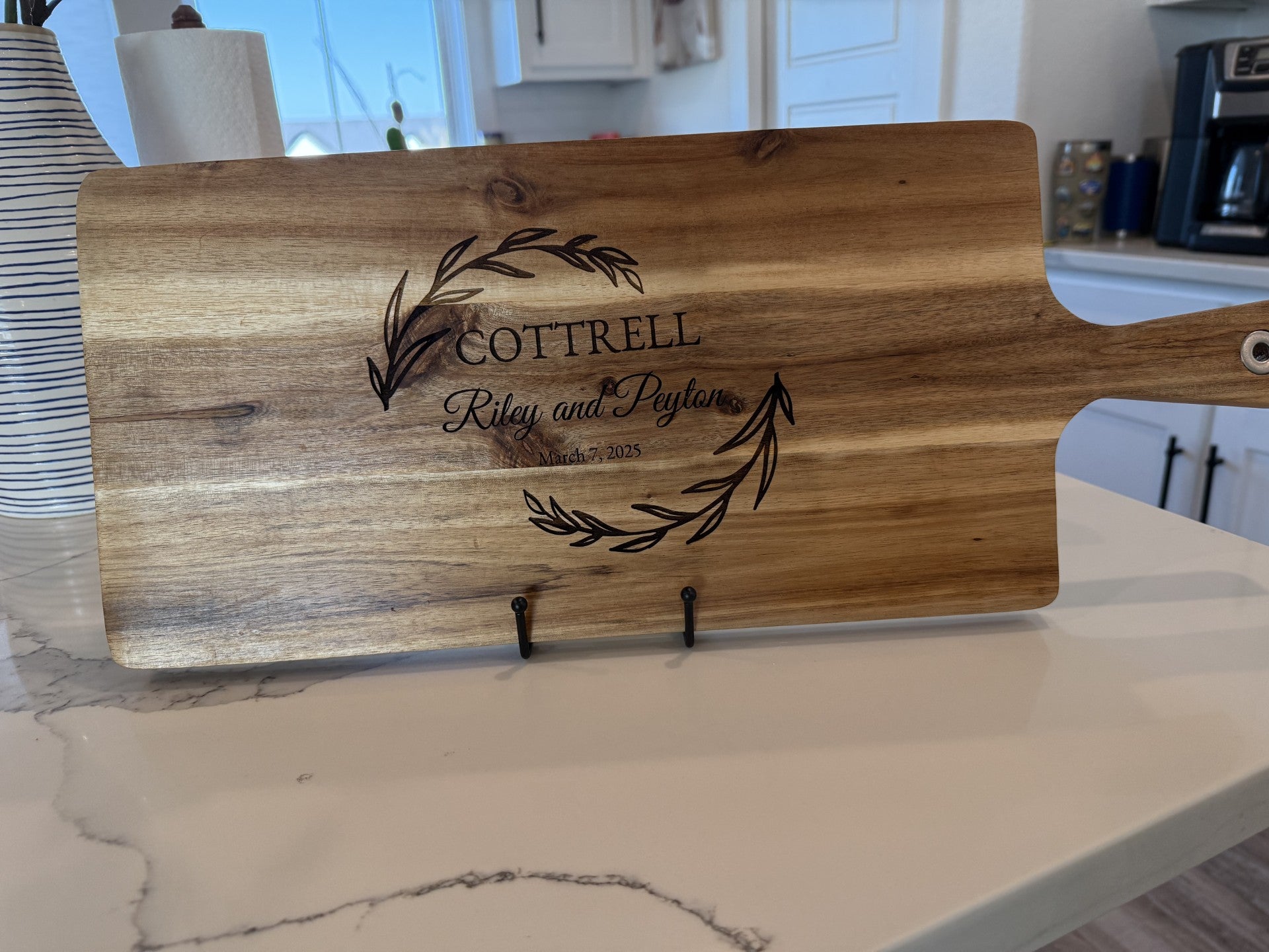 A personalized wooden cutting board, featuring an engraved wreath and Cottrell, Riley and Peyton, Est. March 8, 2023, with a hanging handle is displayed upright on a metal stand against the white marble kitchen counter. Product: Personalized family information cutting board.