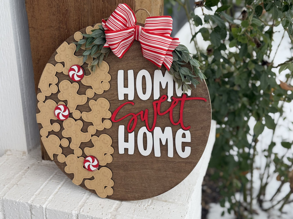 Round wooden sign with the text Home Sweet Home in white and red letters. Decorated with gingerbread shapes, peppermint candies, and a red and white striped bow. Greenery accents are attached at the top. The sign is leaning against a white brick surface outdoors.