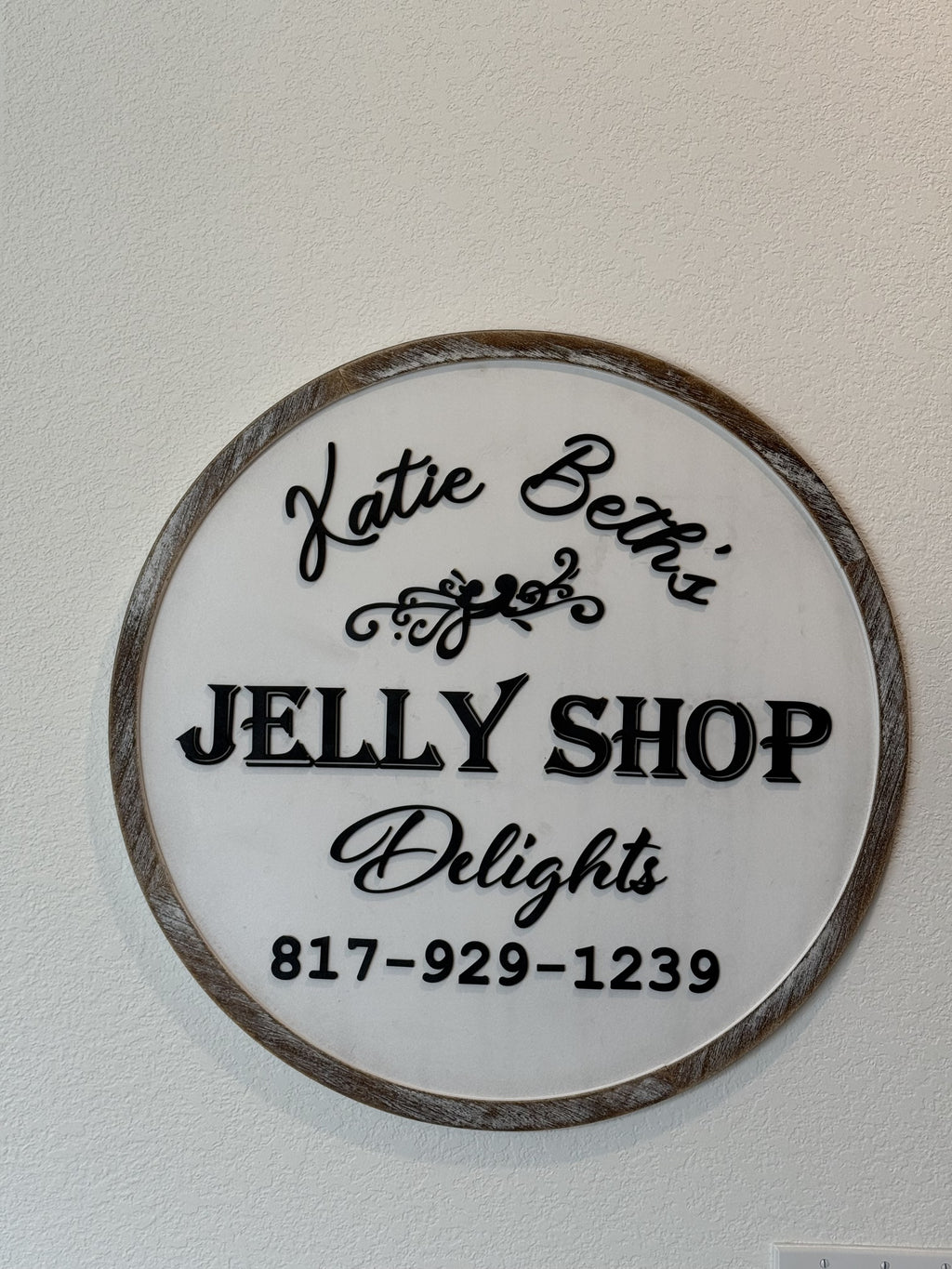 A circular wooden sign with decorative script reads Katie Beth’s Jelly Shop Delights and lists a phone number: 817-929-1239. The sign has a white background and black text, framed by a rustic wooden border, mounted on a pale textured wall.