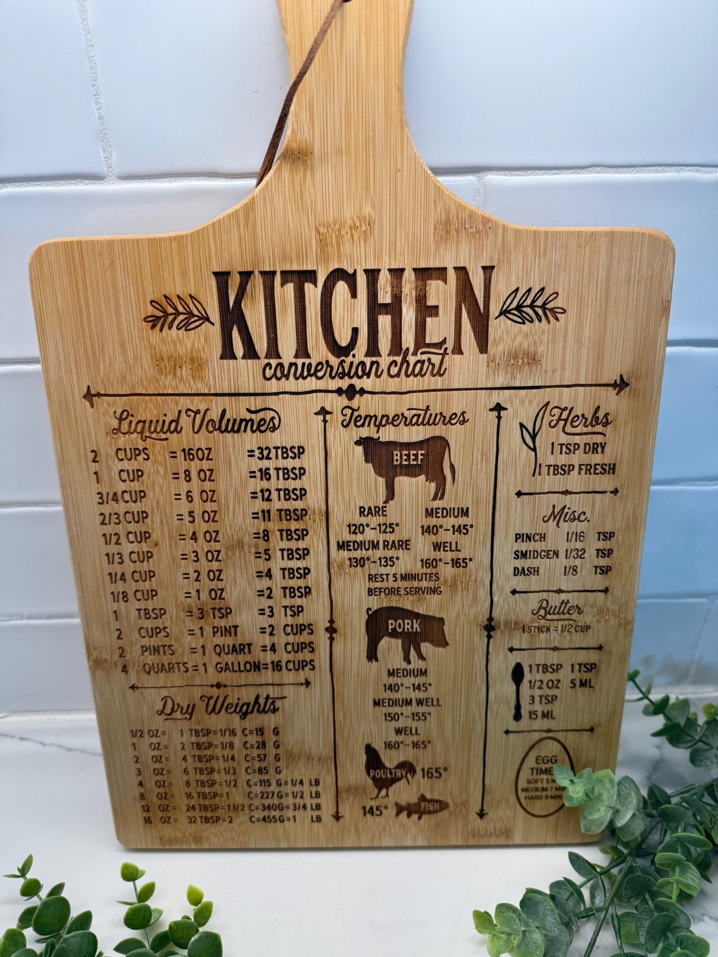 A wooden cutting board with a kitchen conversion chart engraved on it. The chart includes liquid volumes, temperature guidelines for various meats, herb conversions, and dry weights. Decorative greenery is placed at the bottom right corner.