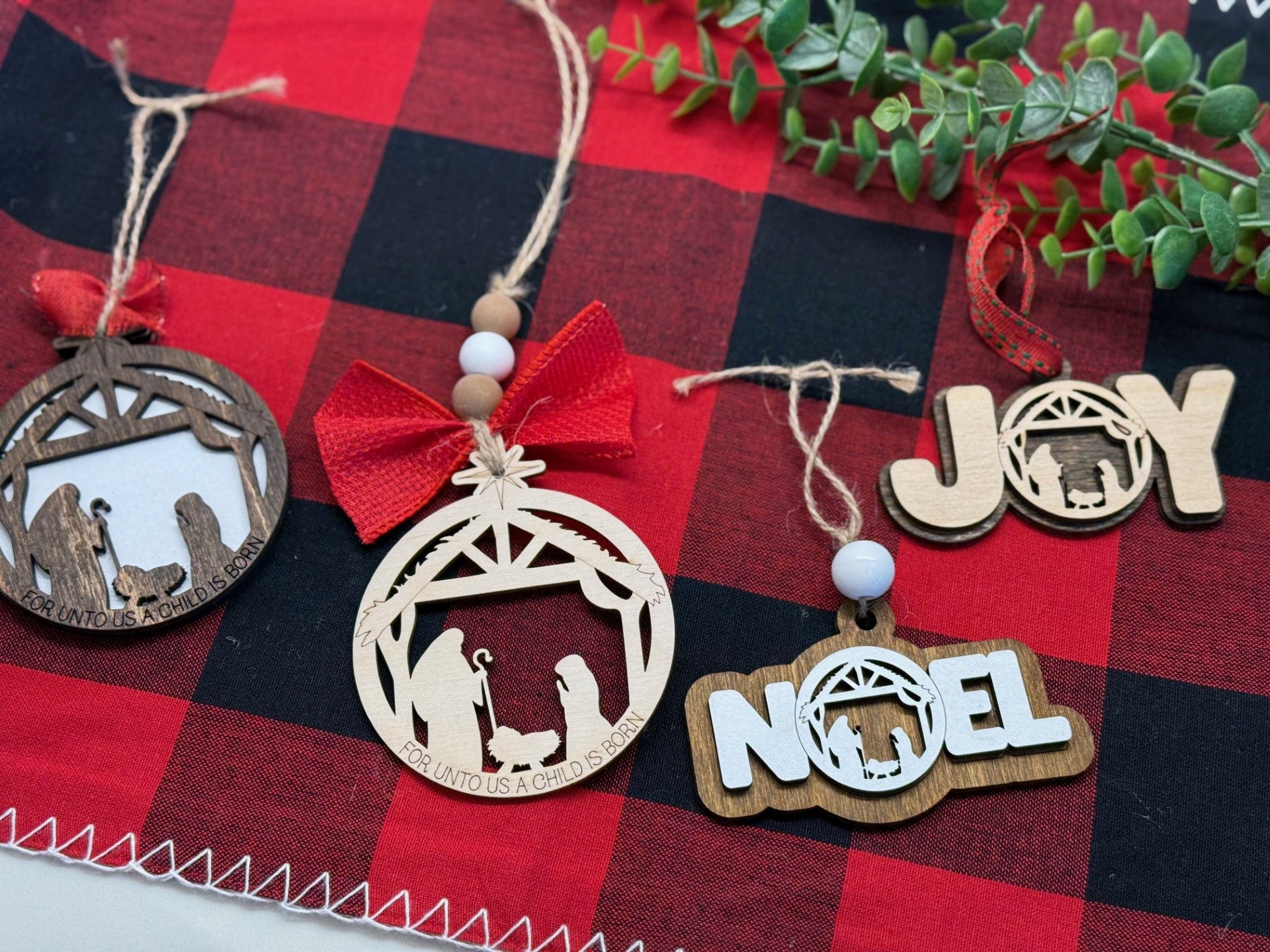 Four rustic Christmas ornaments on a red and black plaid cloth: two wooden nativity scenes, one with a red bow, and two word designs, JOY and NOEL, featuring nativity silhouettes. A sprig of green leaves is in the background.