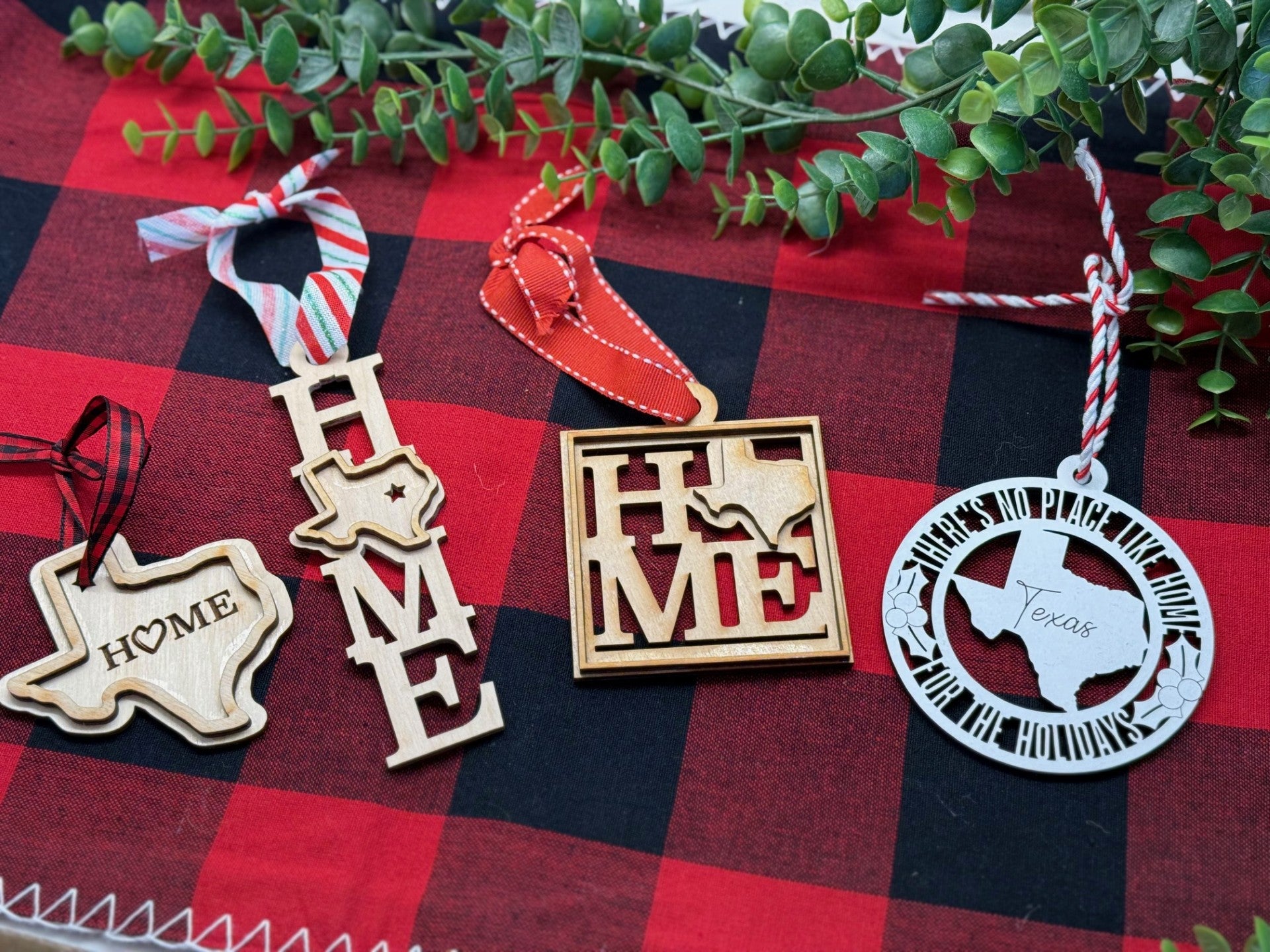Four Texas-themed Christmas ornaments from our collection rest on a red and black checkered cloth. Each wooden design features maps, HOME lettering, decorative ribbons, and is adorned with greenery for a festive touch.