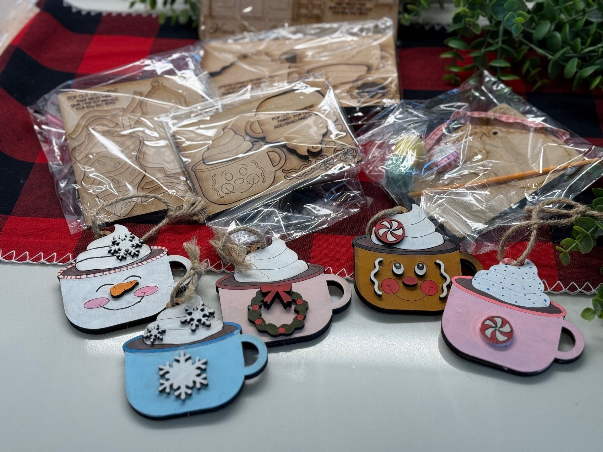 A collection of holiday-themed wooden ornaments shaped like mugs with whipped cream toppings. Each mug is uniquely decorated with elements like snowflakes, cookies, or peppermint candies. Unfinished ornaments are in clear bags in the background. Greenery and a red plaid cloth border the scene.