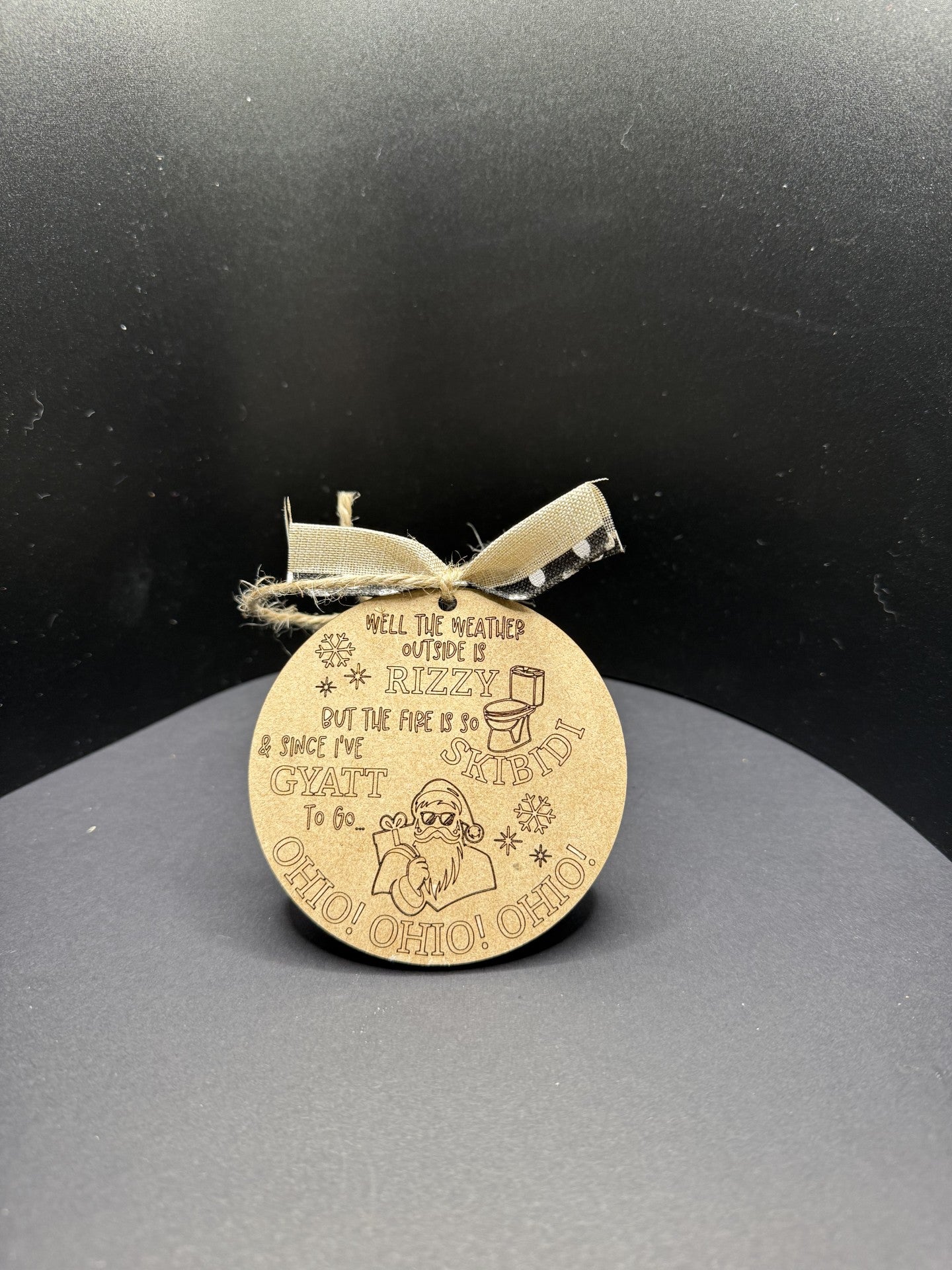 The Rizzy Skibidi Gyatt Ohio Ohio Ohio Christmas Ornament is round and brown with a rustic string. It humorously features Santa and reads, Well the weather outside is RIZZY, but the fire is so GYATT SKIBIDI, adding festive fun with viral internet phrases.