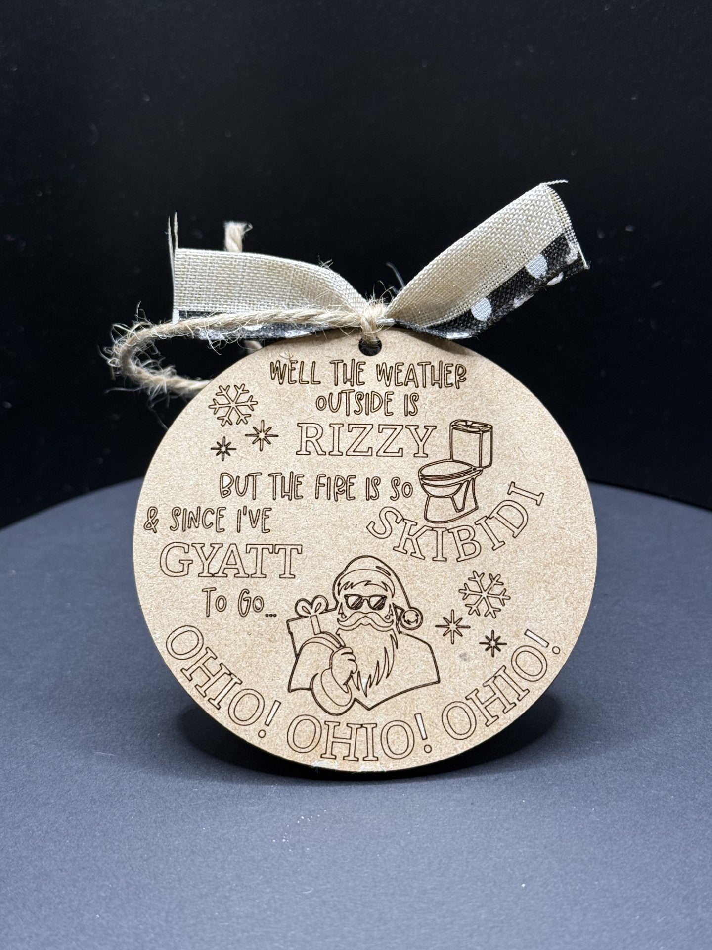 The Rizzy Skibidi Gyatt Ohio Ohio Ohio Christmas Ornament is a round wooden decoration with engraved text and images, featuring a Santa in sunglasses holding a toilet paper roll. The quirky message reads, Well the weather outside is RIZZY, but the fire is so SKIBIDI. It includes a burlap ribbon.