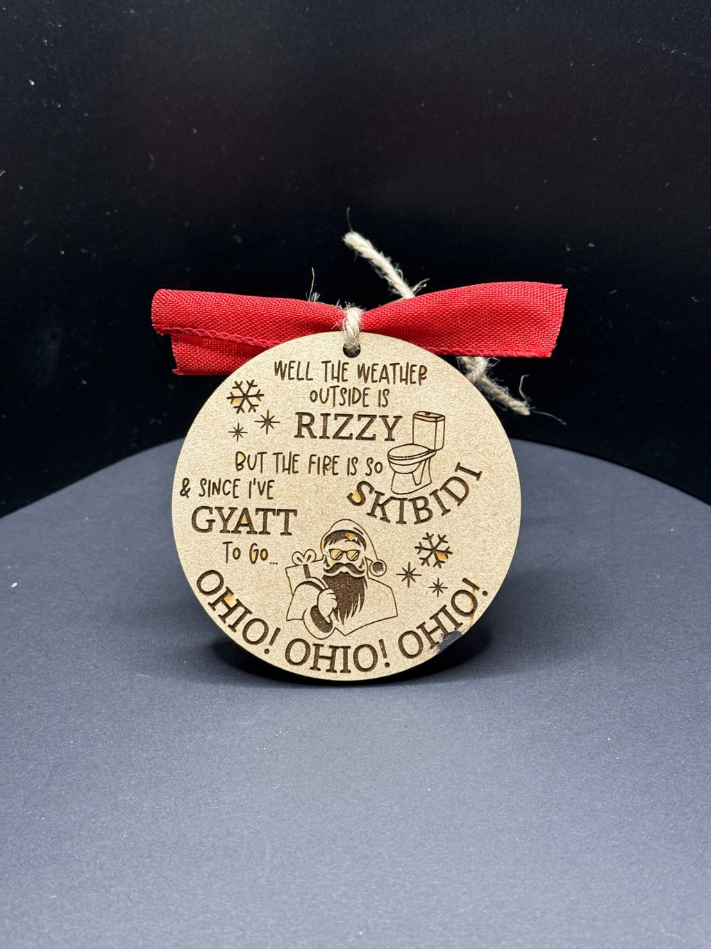 The Rizzy Skibidi Gyatt Ohio Ohio Ohio Christmas Ornament features a round, wood-textured design with engraved text saying, Well, the weather outside is RIZZY, but the fire is so SKIBIDI, & since I’ve got GYATT, along with spirited shouts of OHIO! OHIO! OHIO! and a red ribbon for hanging.