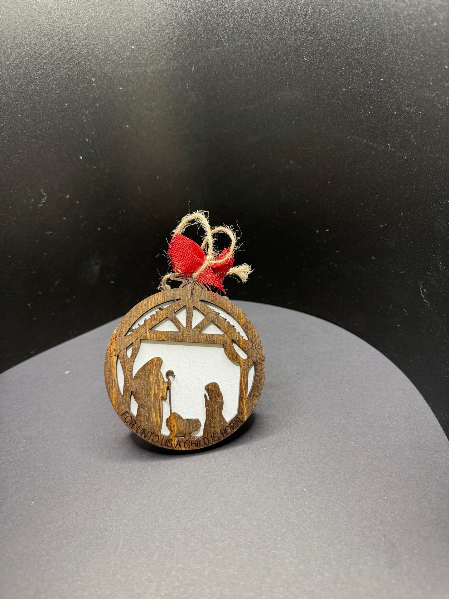 A round, rustic wooden ornament with a Nativity scene cutout, featuring Mary, Joseph, and baby Jesus beneath a stable. The ornament is topped with a red ribbon bow and a twine loop for hanging. The background is a gradient of black and gray.