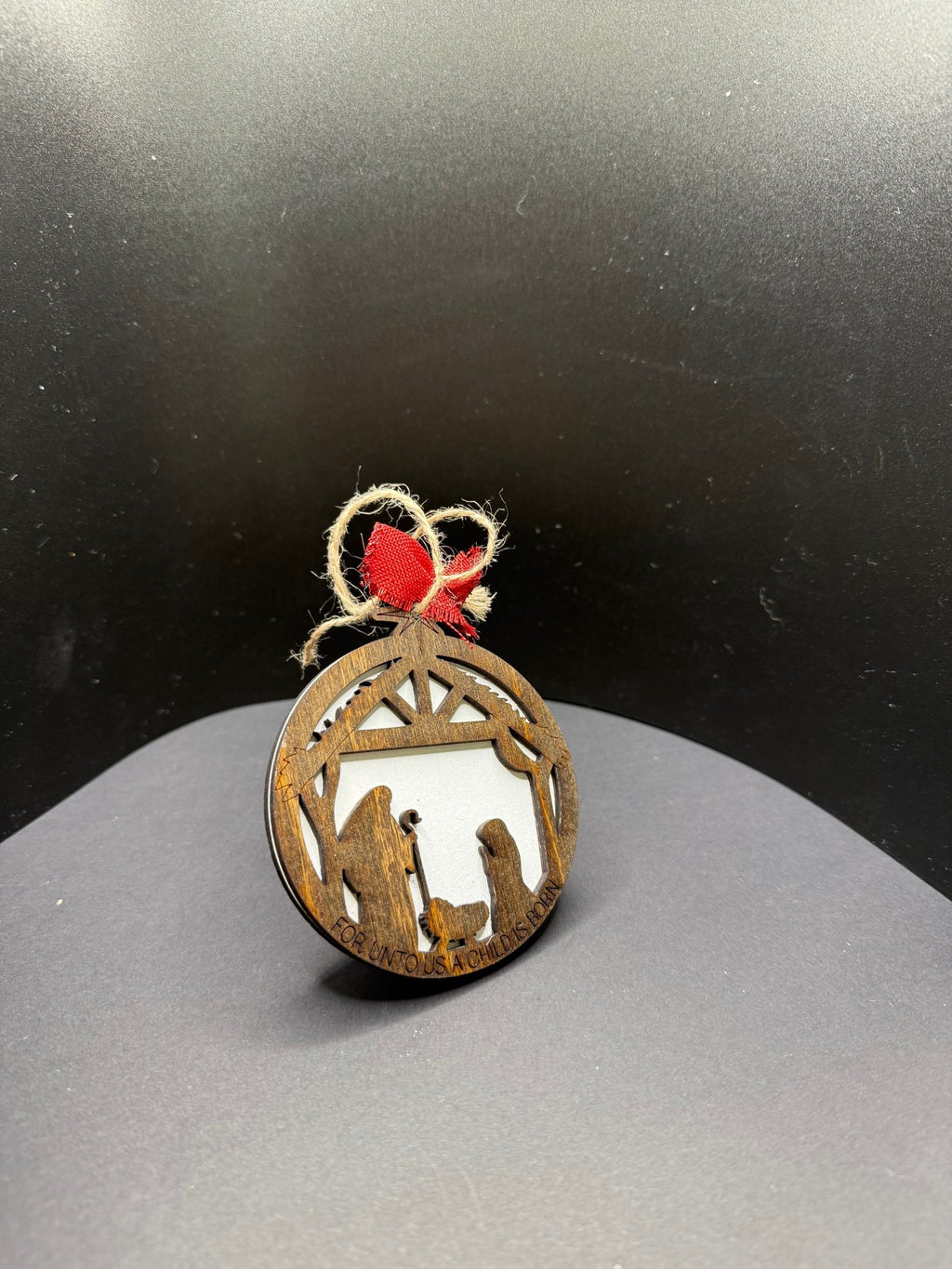 A round, wooden Christmas ornament depicting a nativity scene with a silhouette of Mary, Joseph, and baby Jesus in a stable. Attached is a twine loop with a small red bow for hanging. The ornament rests against a black background.