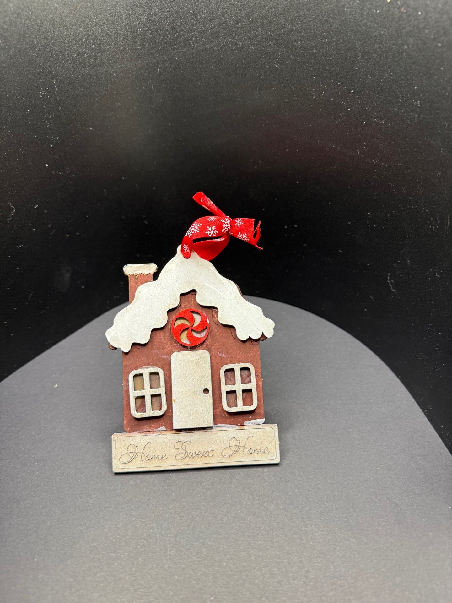 A small, decorative gingerbread house with white frosting details and a peppermint candy above the door. It has a red ribbon with white snowflakes tied to the chimney. The base reads Home Sweet Home. The background is a black curved surface.
