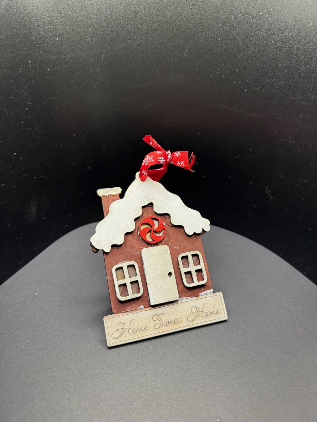 A small, brown wooden ornament shaped like a house with a snow-covered roof and chimney. It has a red and white peppermint candy decoration above the door. A red and white ribbon is attached at the top. The base reads Home Sweet Home. The background is a dark gray gradient.