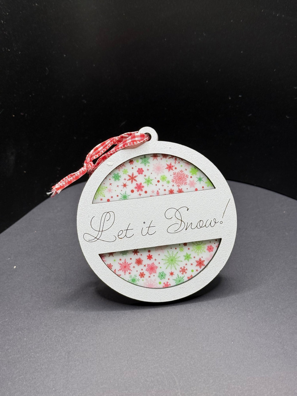 A circular ornament with the phrase Let it Snow! on a white frame. Inside, there is a background of red and green snowflakes. The ornament hangs from a red-and-white checkered ribbon, set against a dark, plain background.