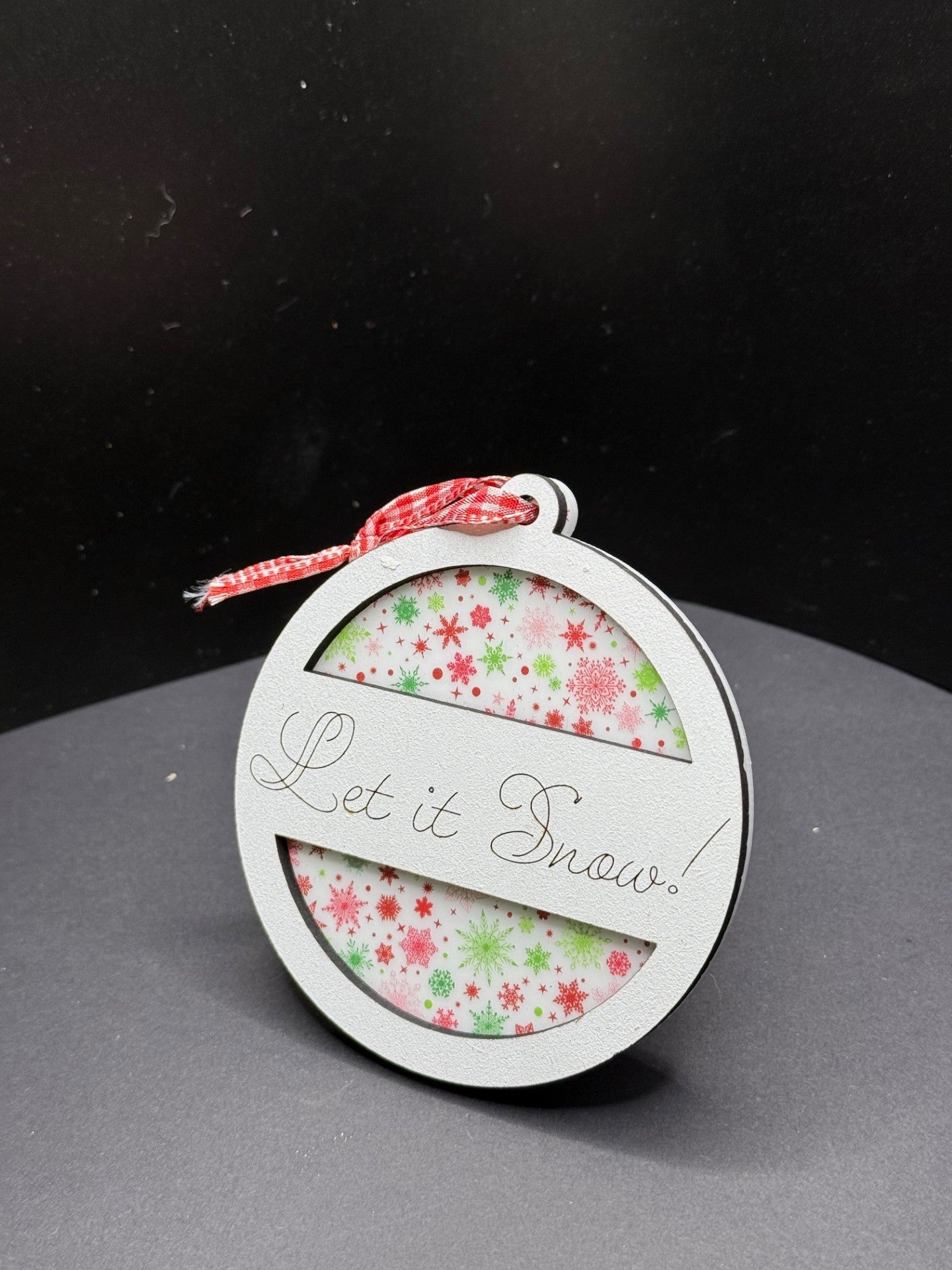 A round ornament with a festive design sits on a dark surface. It features the phrase Let it Snow! on the front. The background inside the ornament is decorated with red and green snowflakes. A small red and white gingham ribbon is tied to the top.