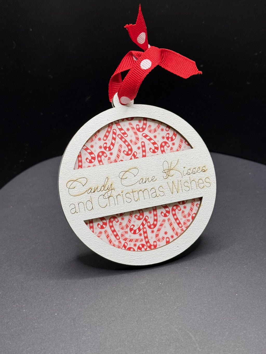 A round, Christmas-themed ornament hangs against a dark backdrop. It features a red candy cane pattern on a white background. A wooden band across the middle reads Candy Cane Kisses and Christmas Wishes in elegant script. A red polka dot ribbon is tied at the top.