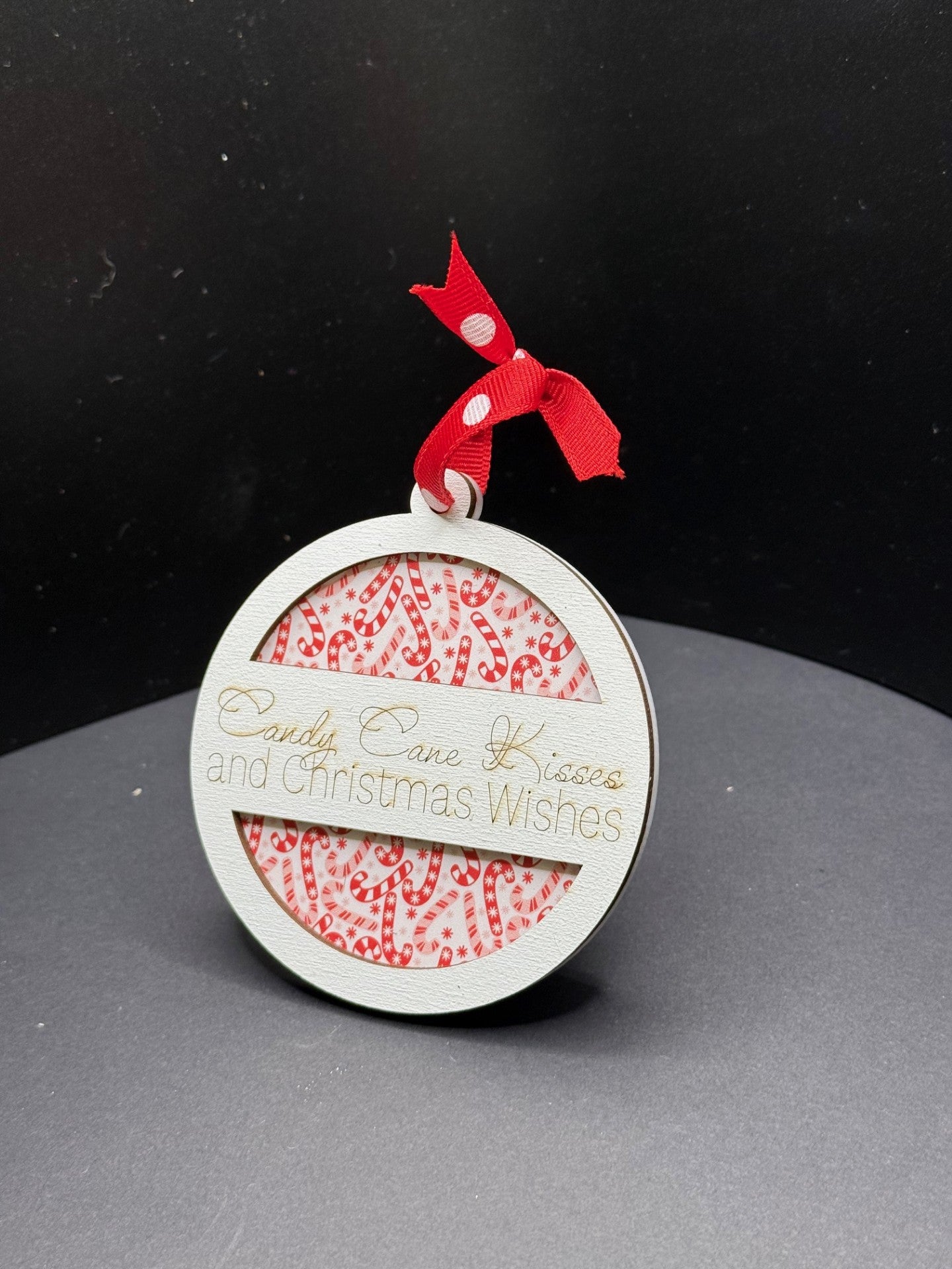 A round Christmas ornament with the text Candy Cane Kisses and Christmas Wishes is displayed. It features a red and white candy cane pattern in the background and is topped with a small red ribbon with white polka dots. The ornament rests on a dark surface.