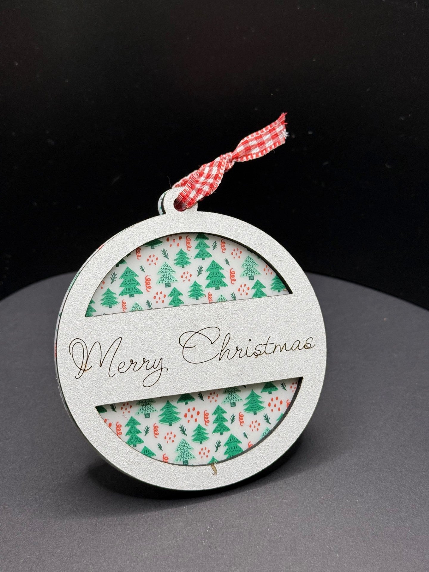 A round Christmas ornament with a white frame, featuring Merry Christmas in elegant script. Inside, there is a festive pattern of green trees and red polka dots. A red and white checkered ribbon is tied at the top. The background is a solid black.