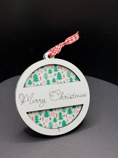 A round Christmas ornament with a white frame, featuring Merry Christmas in elegant script. Inside, there is a festive pattern of green trees and red polka dots. A red and white checkered ribbon is tied at the top. The background is a solid black.