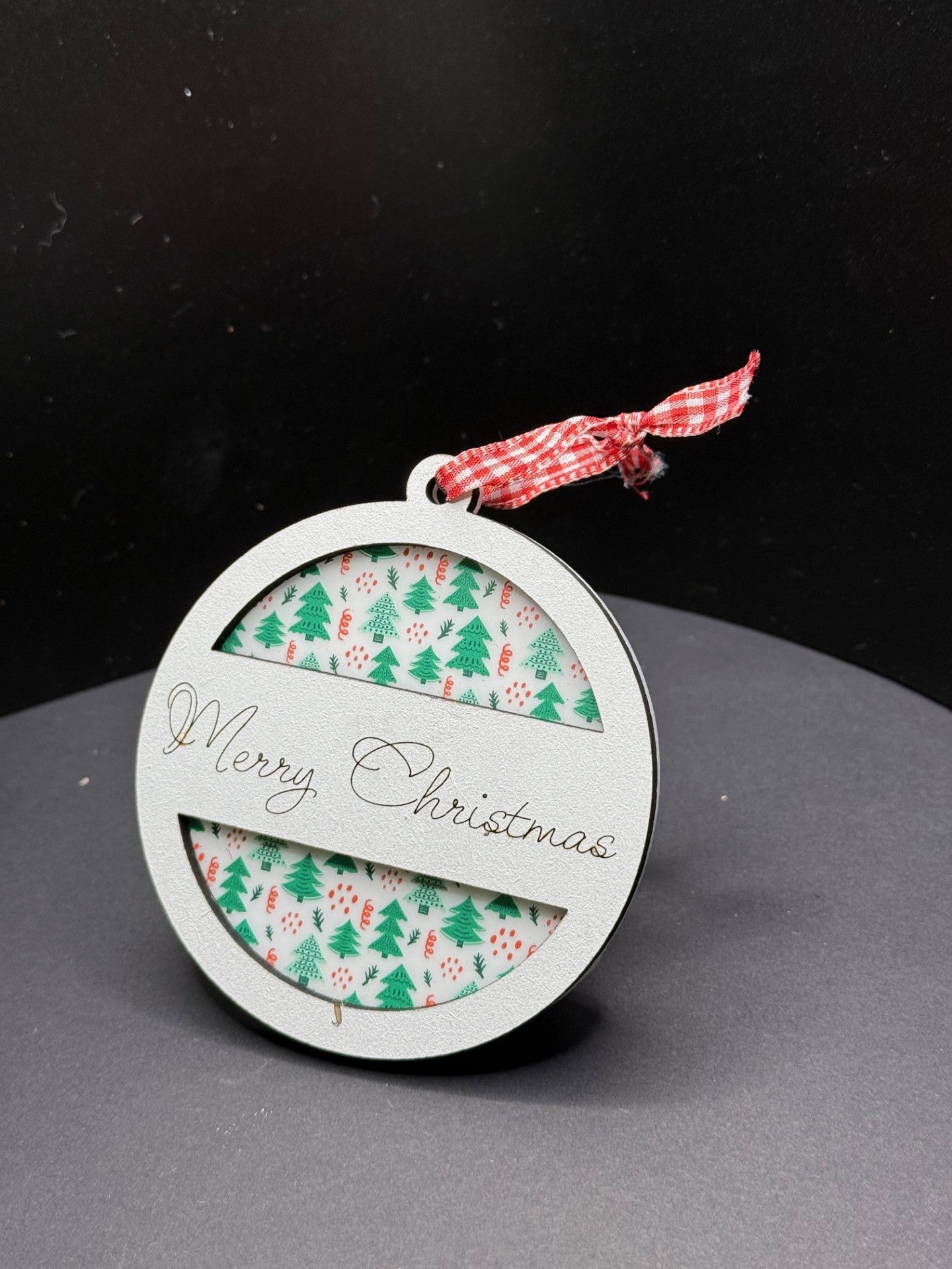 A circular Christmas ornament with a Merry Christmas message. It features a festive pattern of green trees and red decorations. The ornament has a red and white gingham ribbon for hanging, all set against a plain black background.