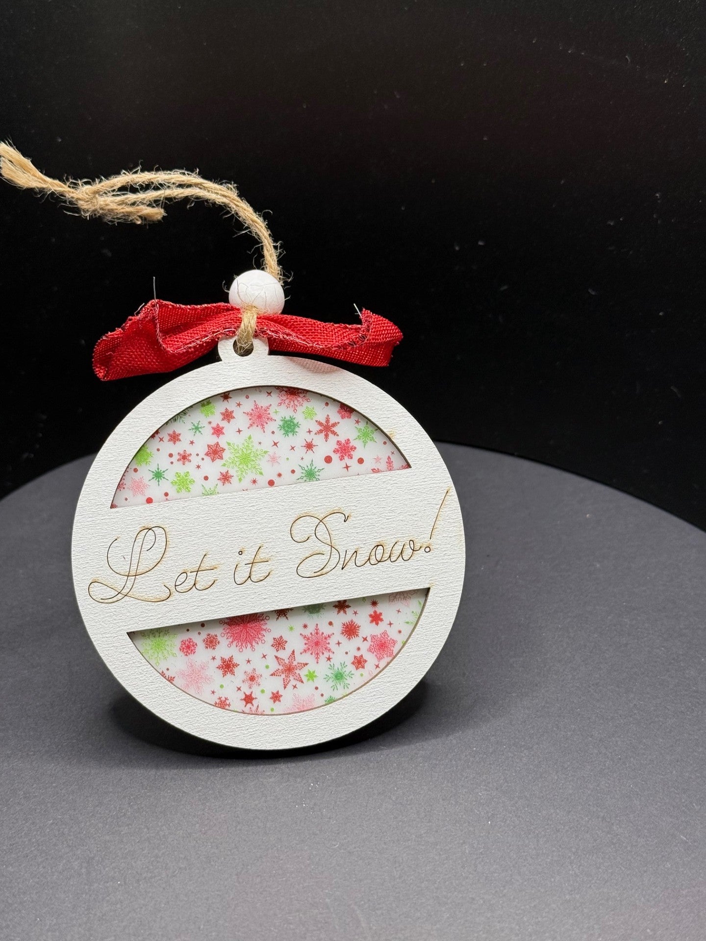 A circular holiday ornament with a red ribbon bow on top, featuring the words Let it Snow! etched on a wooden overlay. The background is patterned with red and green snowflakes. It hangs by a rustic twine loop and is set against a dark backdrop.