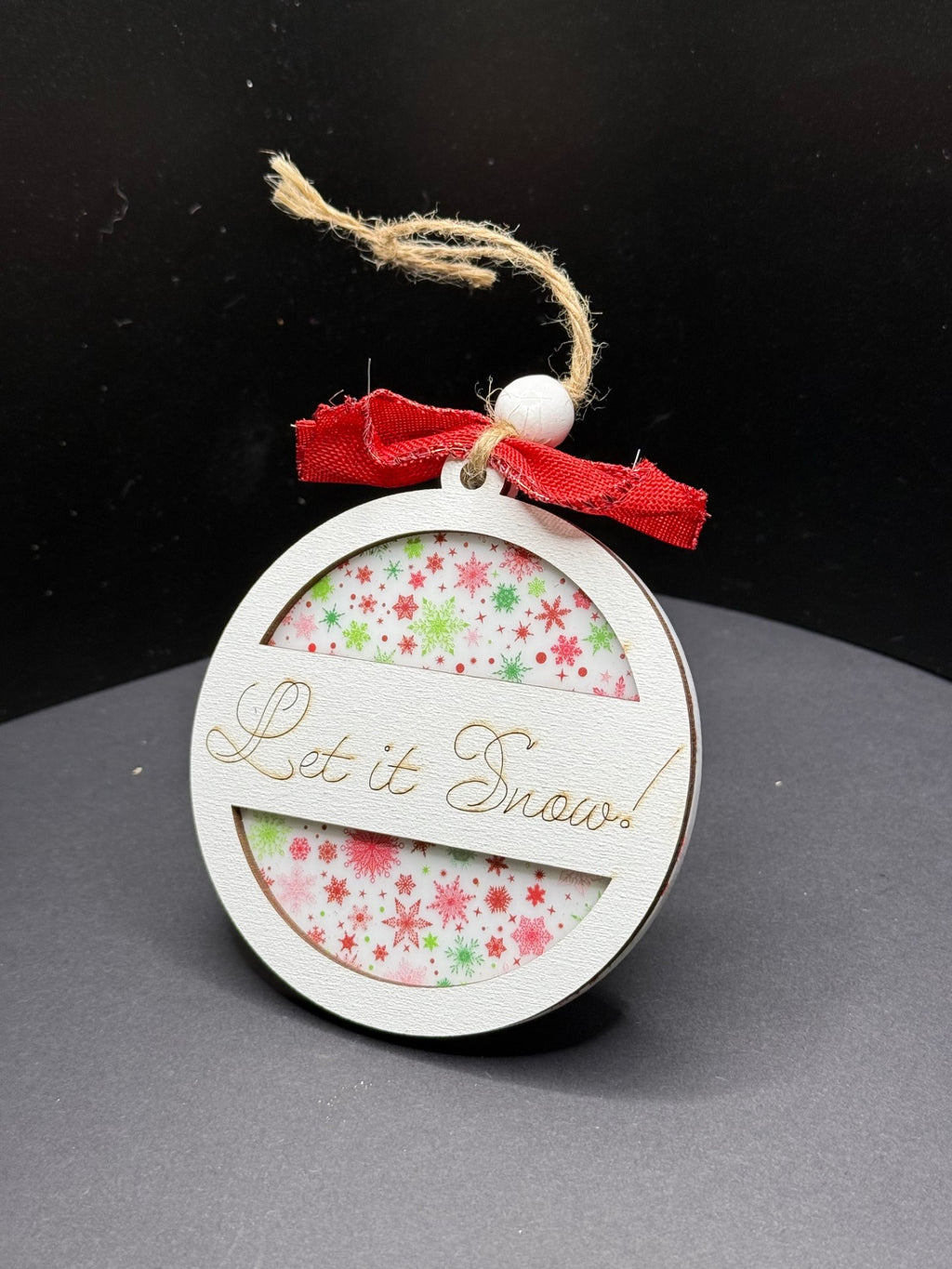 A round wooden ornament with the words Let it Snow! engraved on the front. Inside, theres a red and green snowflake pattern. The ornament is topped with a frayed jute string and a red bow, set against a plain dark background.
