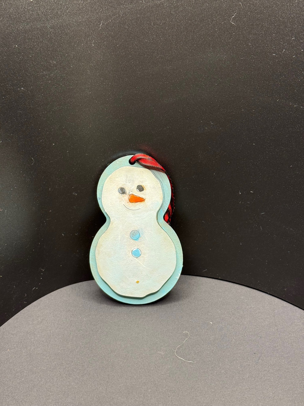A handmade snowman ornament with a light blue body is adorned with a smiling face, an orange carrot nose, and two blue buttons. It hangs against a dark background with a red and black ribbon looped through a hole at the top.