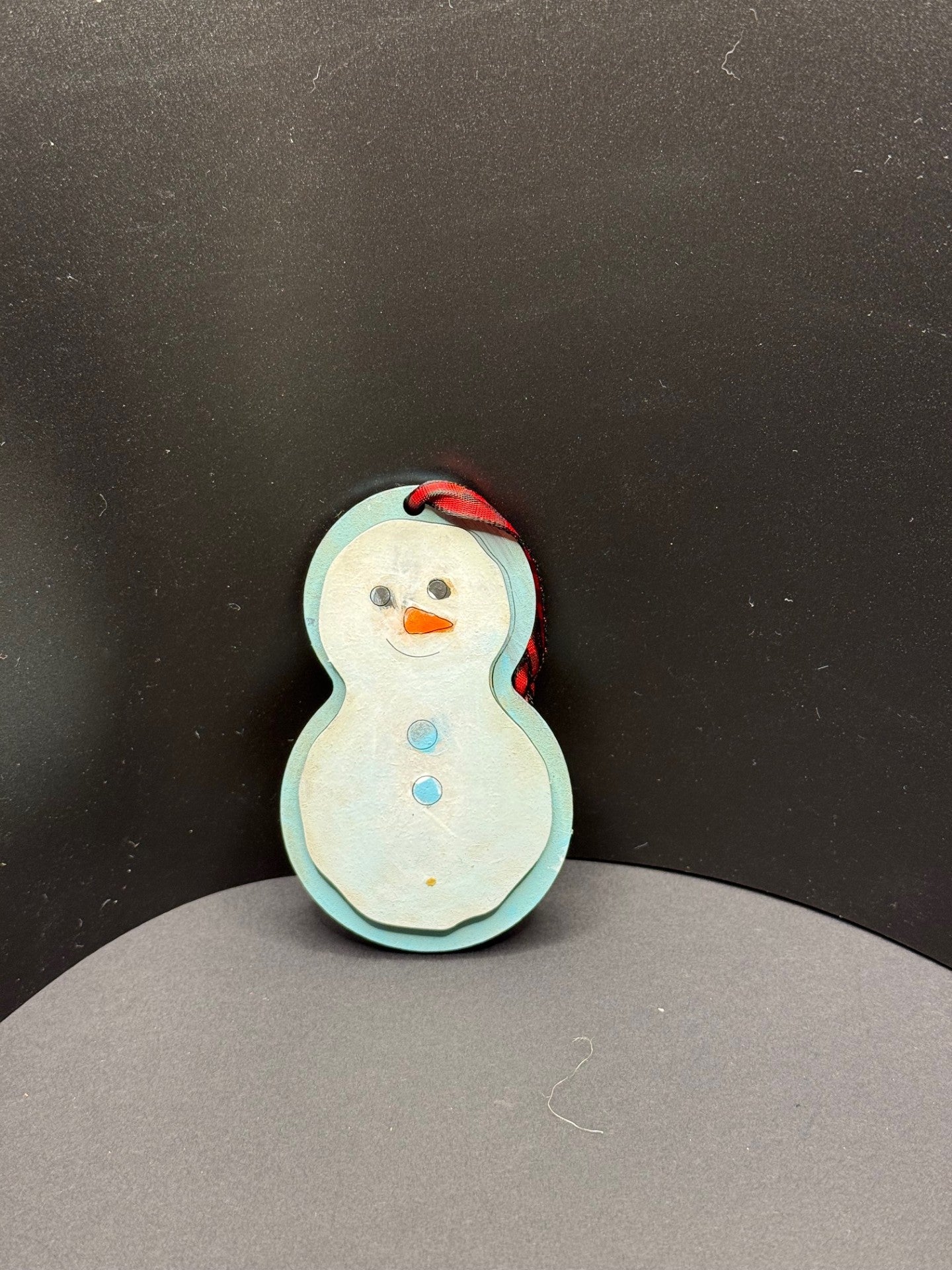 A handmade snowman ornament with a light blue body is adorned with a smiling face, an orange carrot nose, and two blue buttons. It hangs against a dark background with a red and black ribbon looped through a hole at the top.