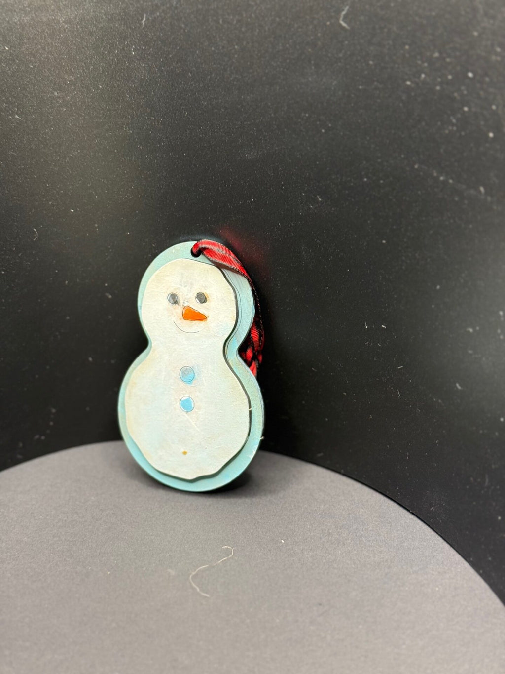 A hand-painted wooden snowman ornament leans against a black background. The snowman features a blue outline, two blue buttons, a carrot-shaped orange nose, and black eyes and mouth. A red and black ribbon is attached to the top.