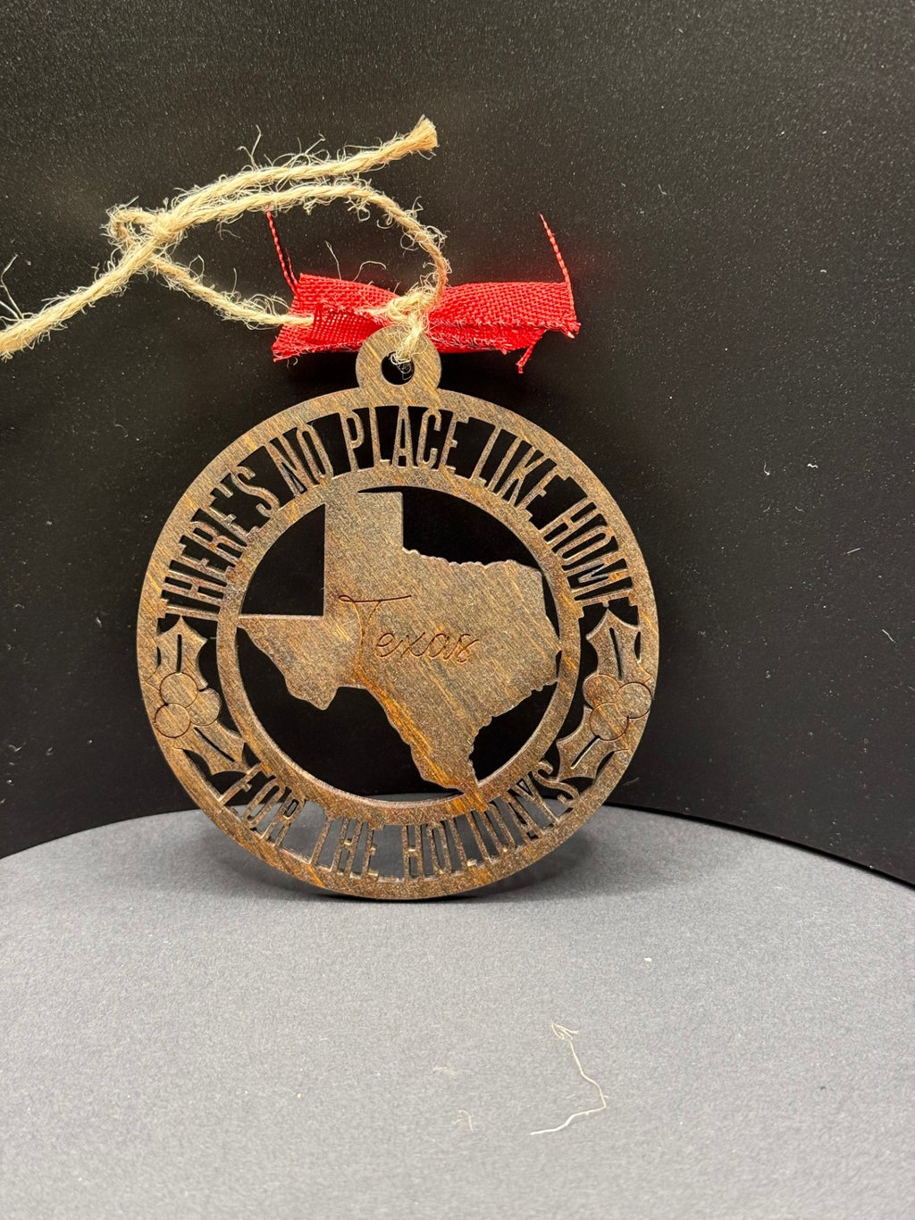 A round, brown ornament hangs on a twine loop with a red ribbon. It features a cutout of Texas with Texas engraved. Surrounding text reads Theres No Place Like Home for the Holidays, with decorative stars and holly. The background is a plain, dark surface.