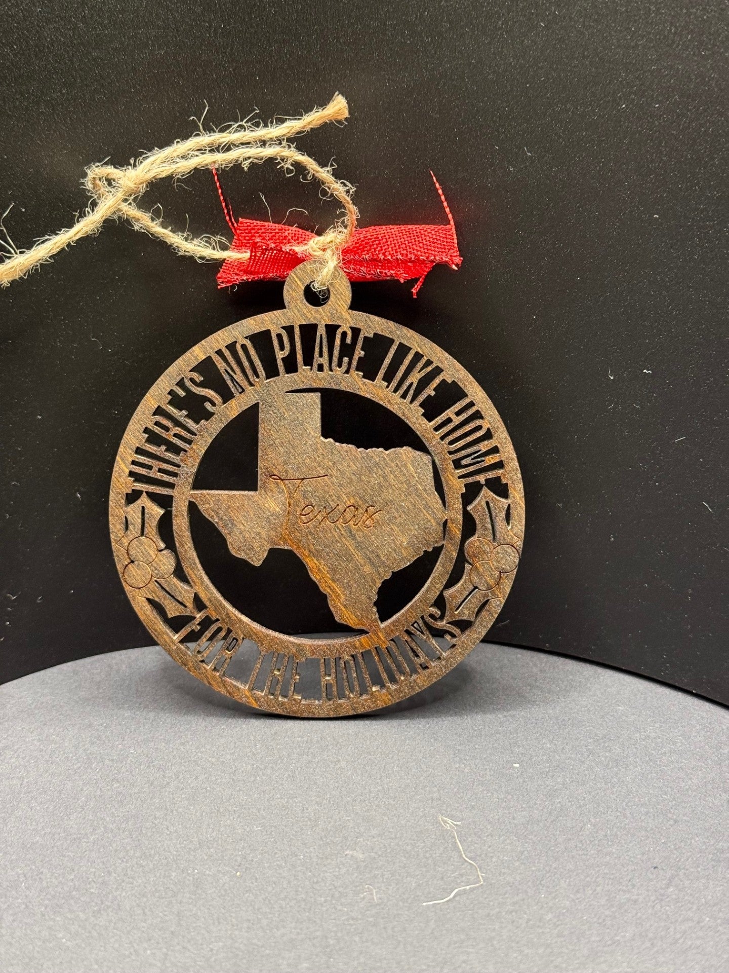 A round, brown ornament hangs on a twine loop with a red ribbon. It features a cutout of Texas with Texas engraved. Surrounding text reads Theres No Place Like Home for the Holidays, with decorative stars and holly. The background is a plain, dark surface.