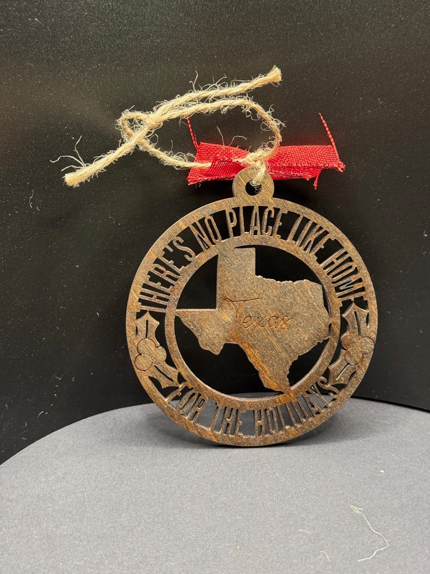 A round wooden ornament with a cutout of Texas in the center, featuring the word Texas. It is adorned with stars and heart motifs. The text around the edge reads Theres No Place Like Home For The Holidays. The ornament hangs from a twine loop with a red bow.