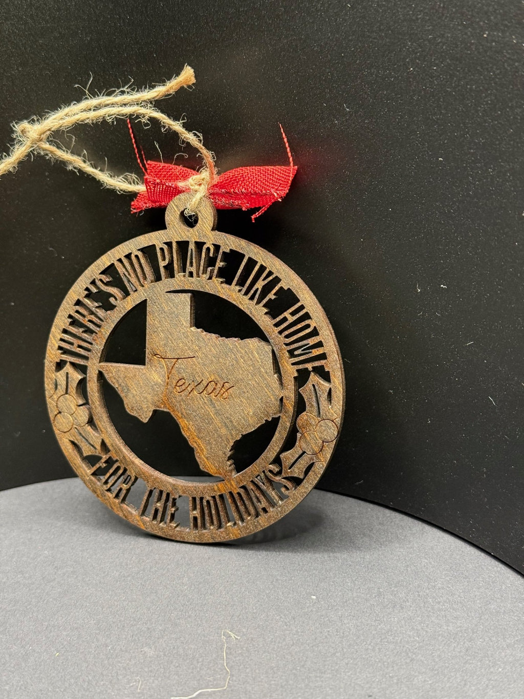 A circular wooden ornament with a cutout design featuring the shape of Texas in the center, engraved with the word Texas. Surrounding the shape, the text reads, THERES NO PLACE LIKE HOME FOR THE HOLIDAYS. The ornament is tied with a twine loop and a small red ribbon.