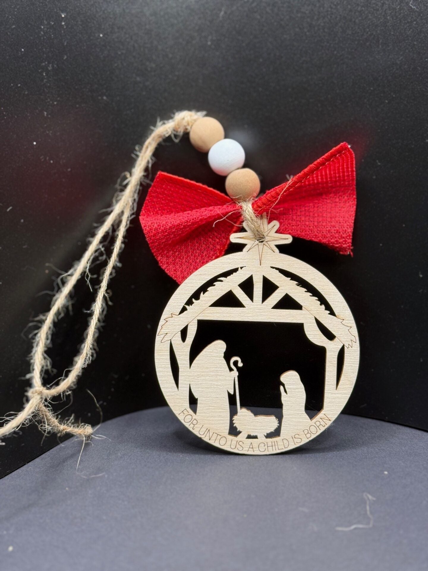 A wooden Christmas ornament features a nativity scene with Mary and Joseph. Above them is a star-topped arch. Below, the text reads FOR UNTO US A CHILD IS BORN. The ornament is adorned with a red bow and beige beads. A jute string is attached for hanging.