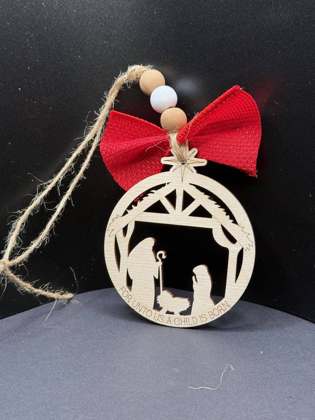 A round wooden Christmas ornament featuring a Nativity scene with Mary, Joseph, and baby Jesus in a manger under a stable. The top has a red fabric bow, a star, and a jute string with wooden beads. The text FOR UNTO US A CHILD IS BORN is engraved at the bottom.