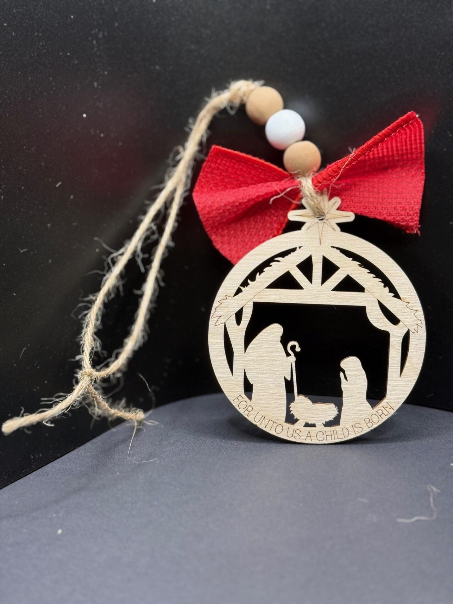 A round wooden ornament depicts a nativity scene with Mary, Joseph, and baby Jesus in a manger. The top is adorned with a red bow and wooden beads. Below the scene, the inscription reads, For unto us a child is born. The ornament hangs by a rustic twine loop.