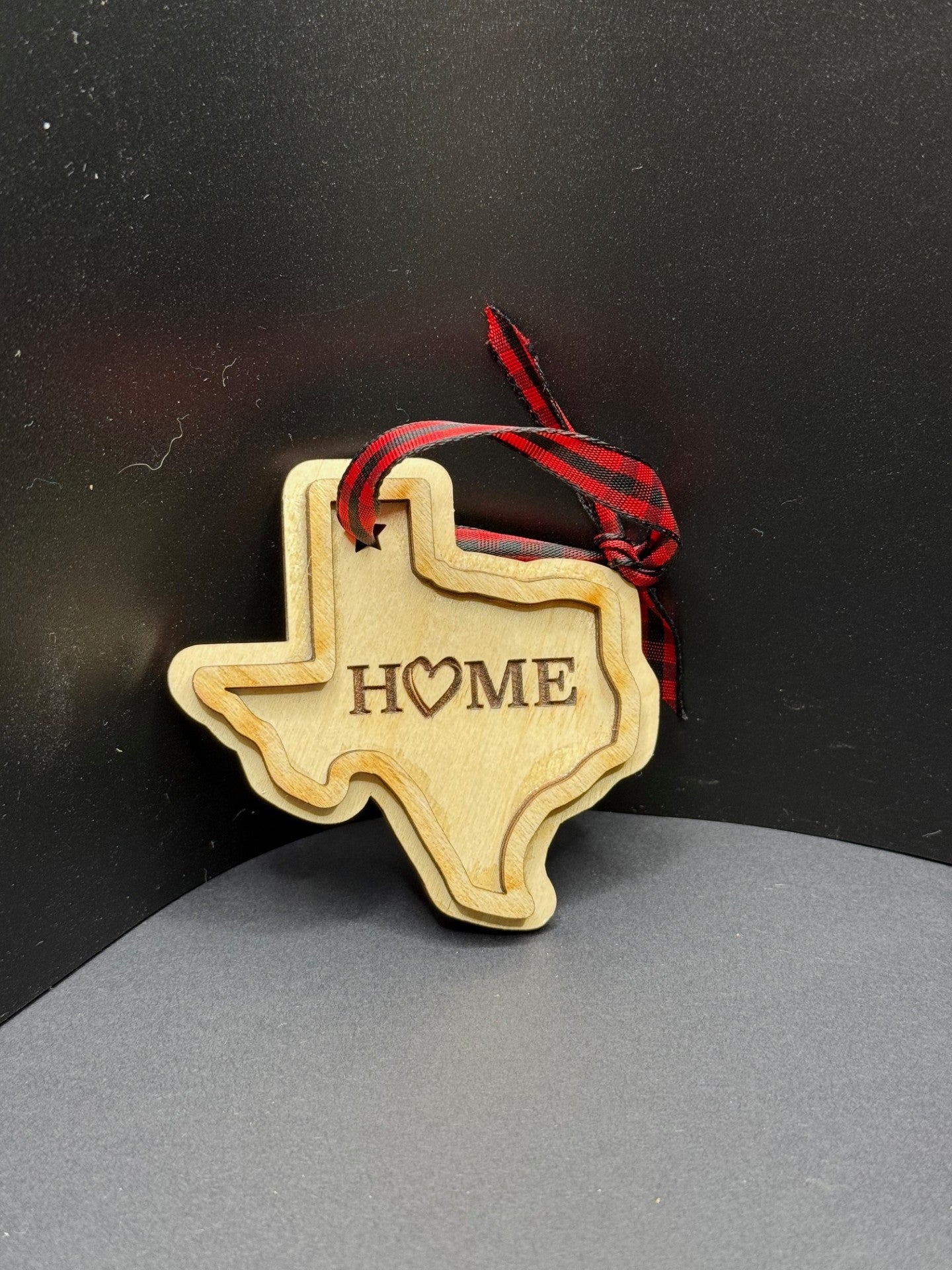 A wooden ornament in the shape of Texas hangs against a dark backdrop. The word HOME is engraved in the center, with a heart symbol replacing the O. A red and black plaid ribbon is attached for hanging.