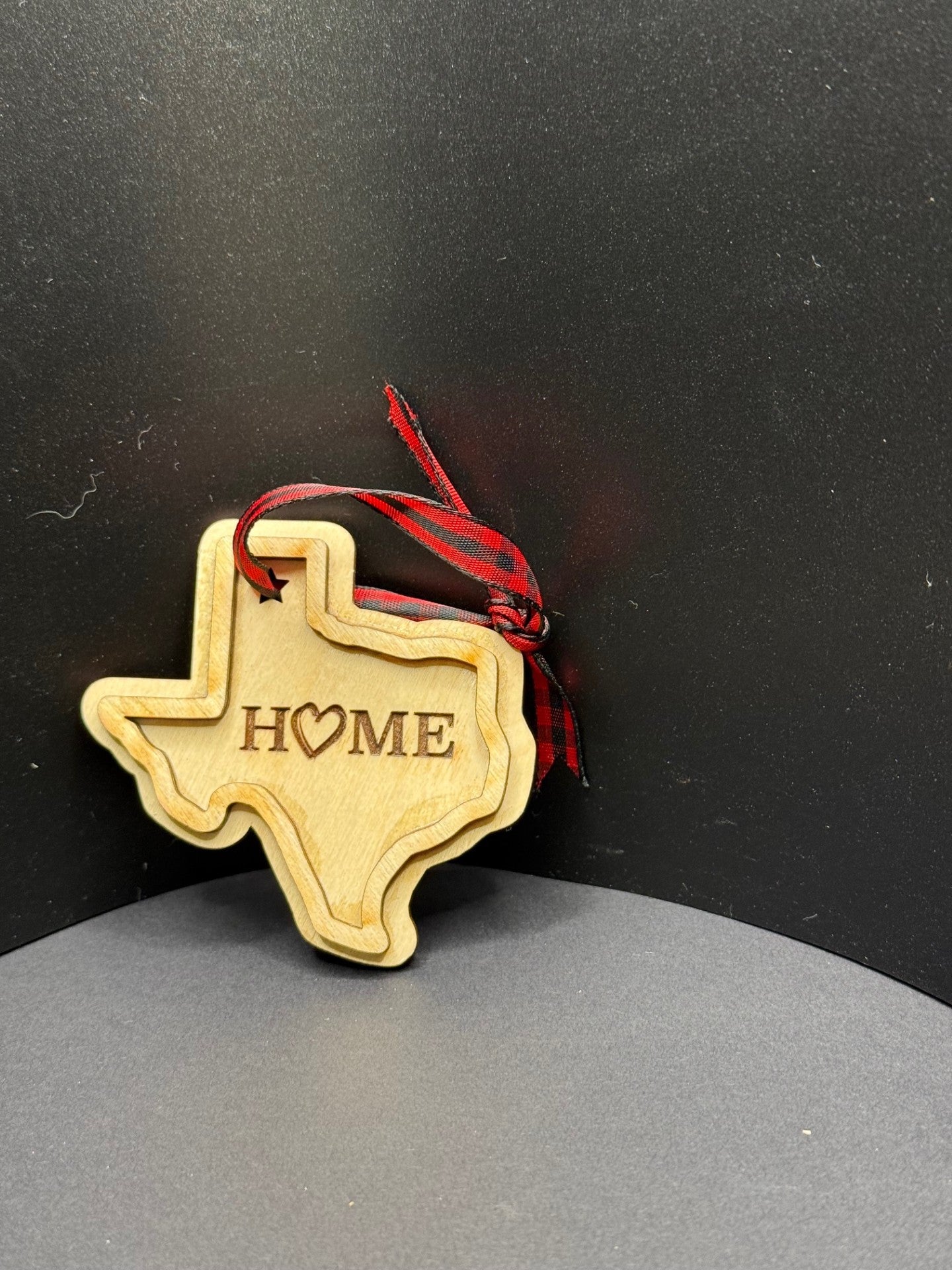 A wooden ornament shaped like the state of Texas with the word HOME engraved on it. The O is represented by a heart. It is adorned with a small red and black checkered ribbon for hanging. The background is a simple, dark surface.