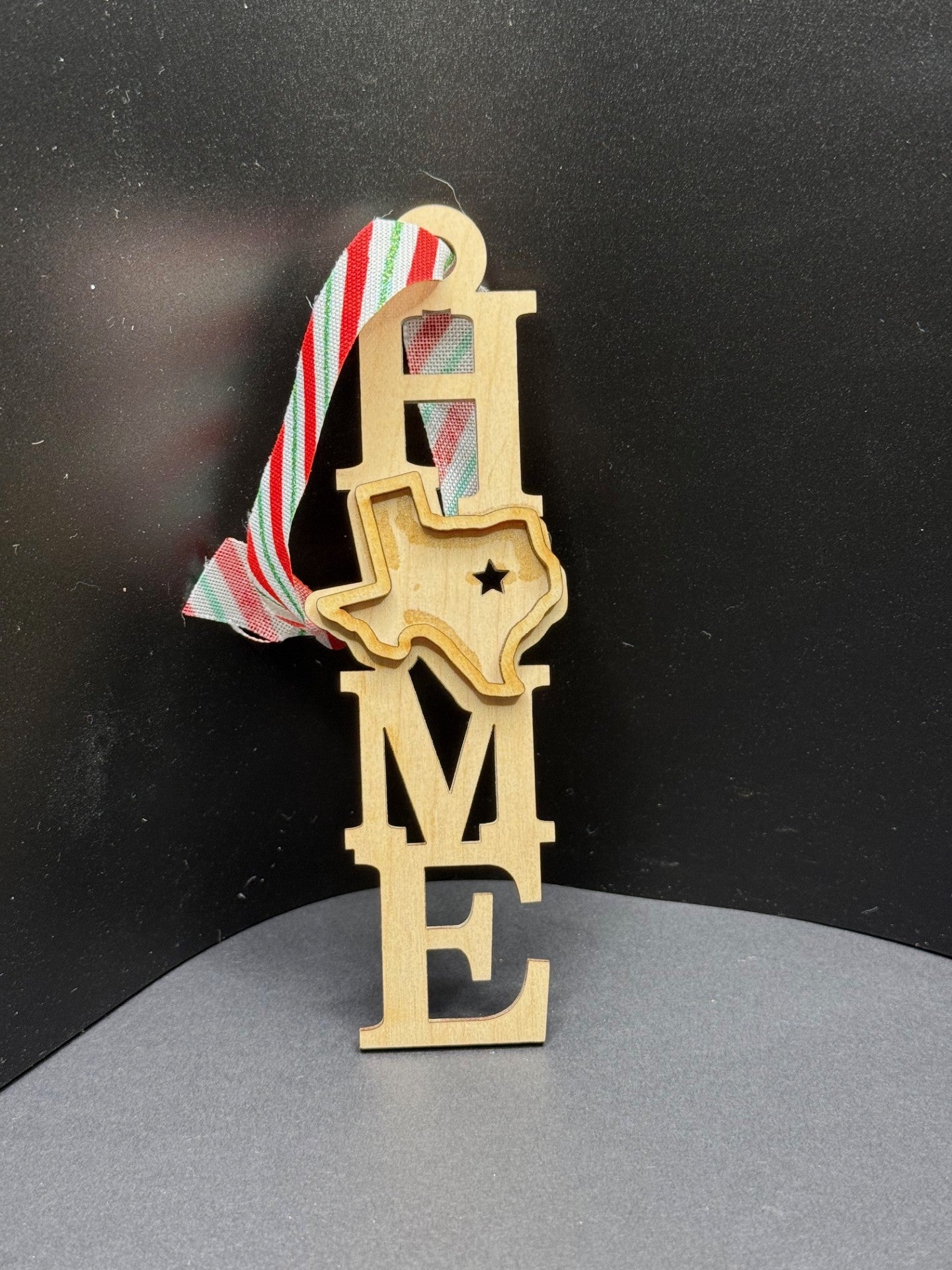 Wooden ornament spelling HOME vertically, with the letter O replaced by a cutout of Texas featuring a star. A red, white, and green striped ribbon is attached for hanging. The ornament is set against a dark background.