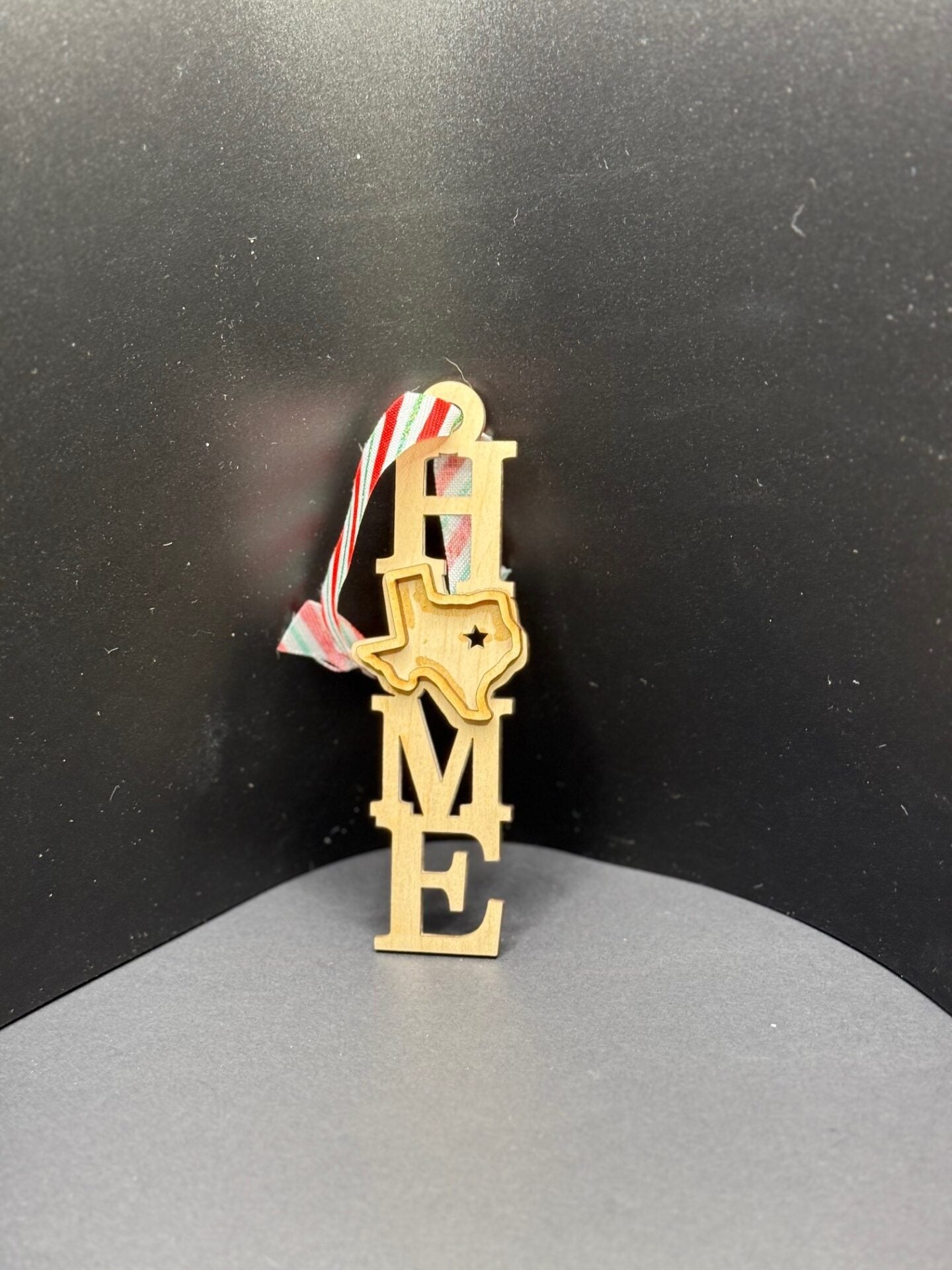 A vertical ornament featuring the word HOME in wooden letters. The letter O is replaced by the shape of the state of Texas, with a star marking a location. It hangs from a red, white, and green striped ribbon. The background is plain black.