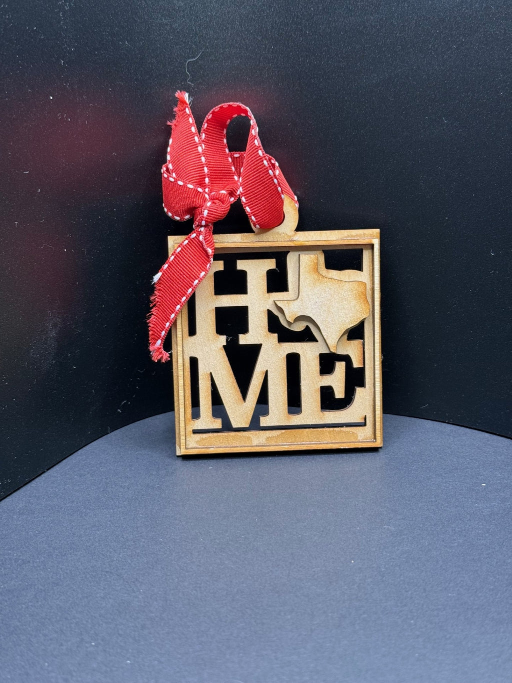Wooden ornament featuring the word HOME with the O replaced by an outline of Texas. The ornament is square with a red ribbon tied at the top. The background is a plain dark surface.