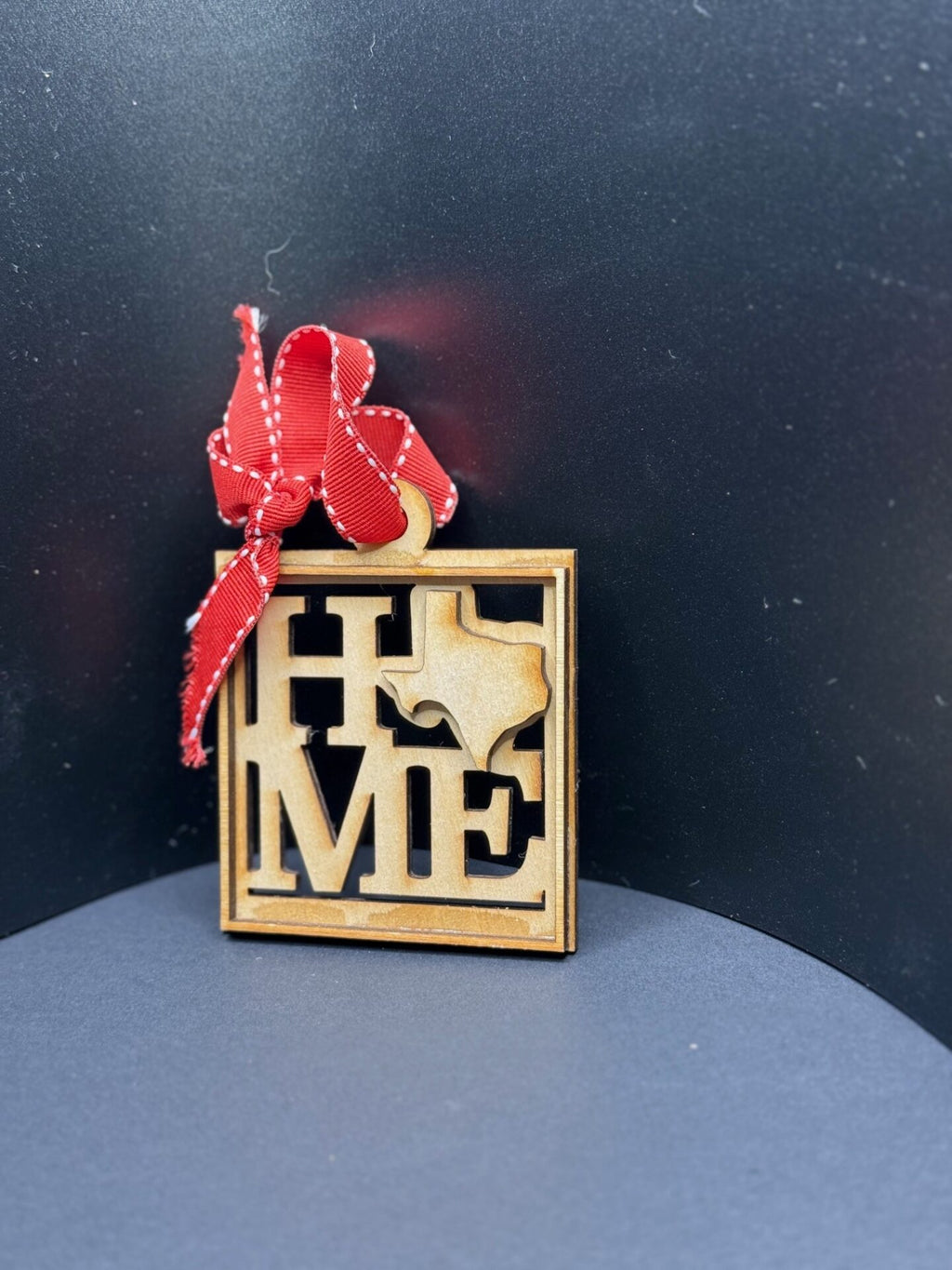A wooden ornament featuring the word HOME with the O replaced by a cutout of Texas. A red ribbon with white stitching is tied in a bow at the top. The background is dark, creating a contrast that highlights the details of the ornament.
