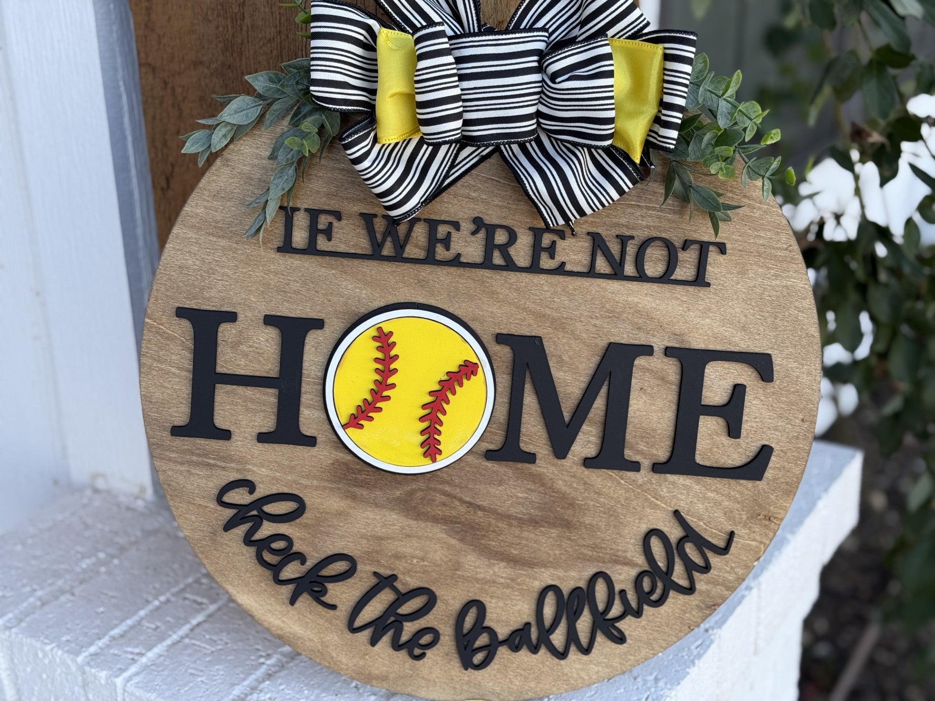 A wooden sign with the text If were not home, check the ballfield. The word home is highlighted with a yellow softball for the letter O. It is embellished with a black and white striped bow with yellow accents and has greenery attached on top.