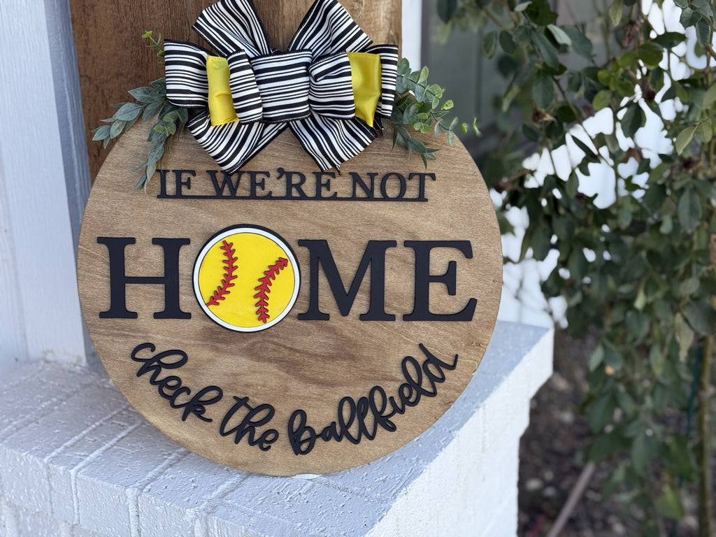 A wooden round sign is displayed on a ledge. It reads, If were not home, check the ballfield, with a yellow softball illustration in the word home. A black and white striped bow with yellow ribbons and green leaves adorns the top. Foliage is in the background.
